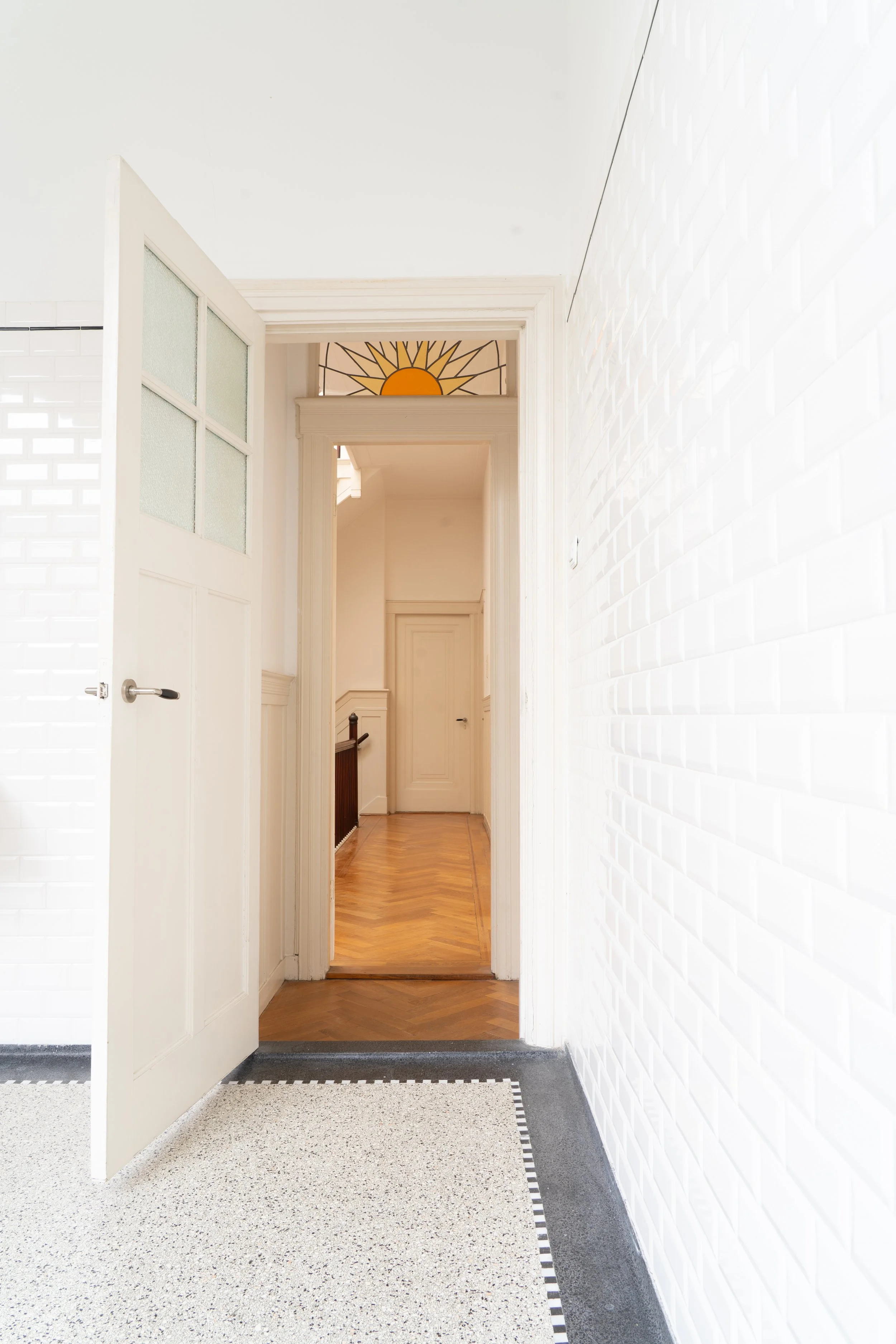 Interieur van een wit gekaleide gang met een open deur naar een ruimte met houten vloer en een plafond met geel en zwart decoratief glasraam.