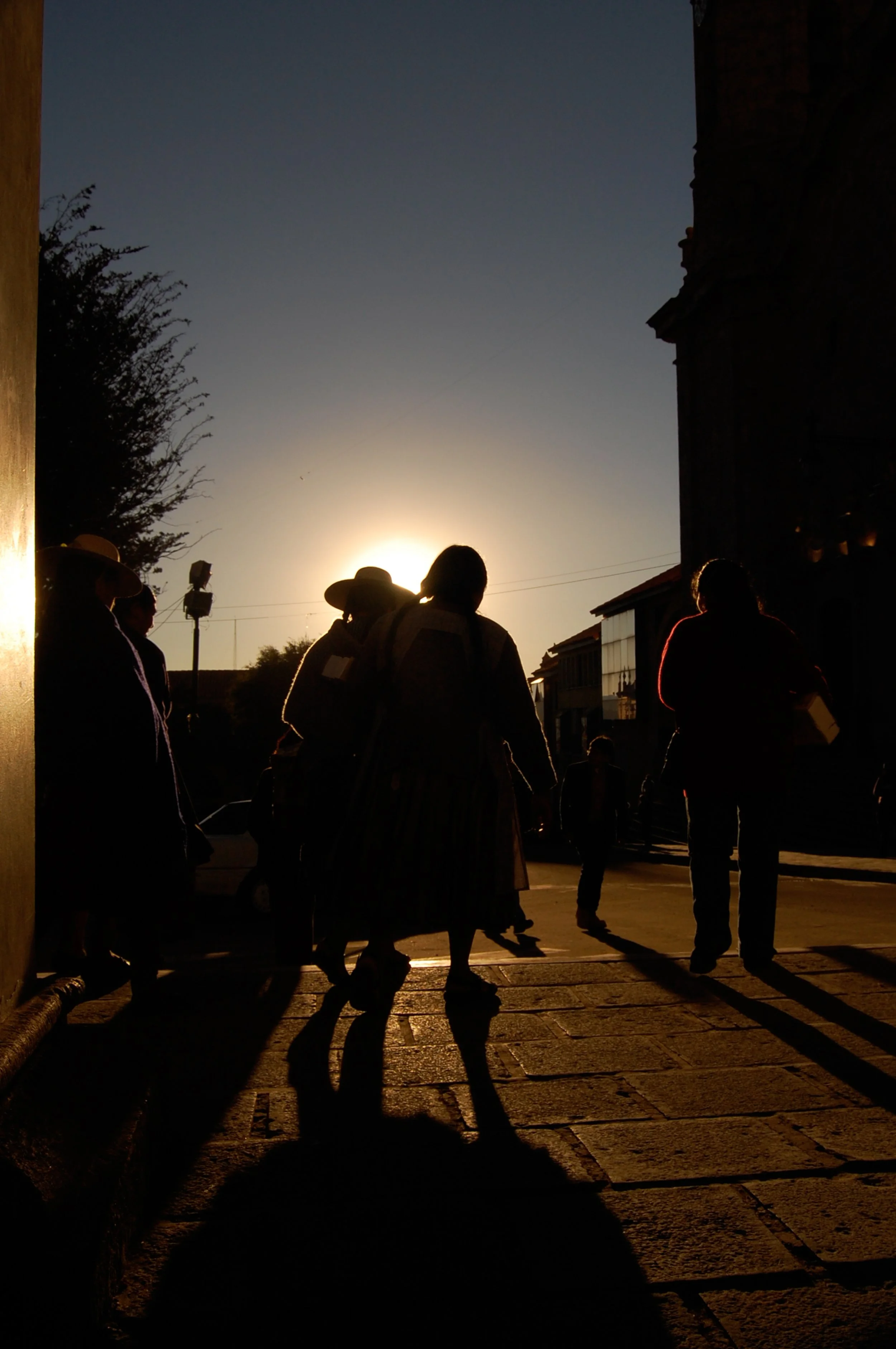 Silhouetten van mensen die in de avond of vroege ochtend over straat lopen met de zon op de achtergrond, gebouwen en een boom zichtbaar, schemerachtige sfeer.