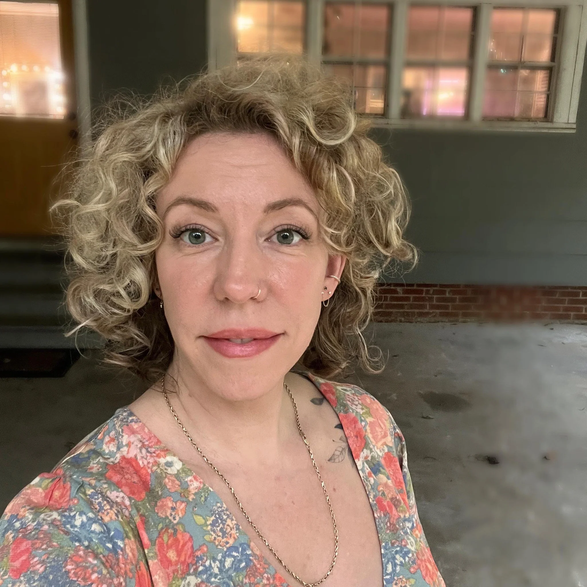 A woman with curly blond hair and blue eyes, wearing a floral dress, gold jewelry, and face piercings, standing in front of a brick building with windows.