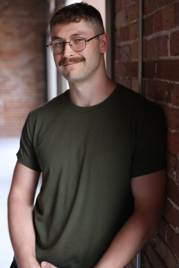 A man with glasses and a mustache is standing against a brick wall, wearing a dark green t-shirt, smiling slightly.