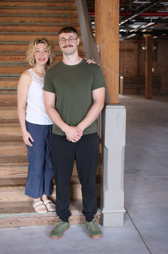 A man and a woman standing near a wooden staircase in a rustic, indoor space. The woman has curly blonde hair, is wearing a white sleeveless top and striped pants. The man has short hair, a mustache, glasses, and is wearing a green t-shirt and black pants.