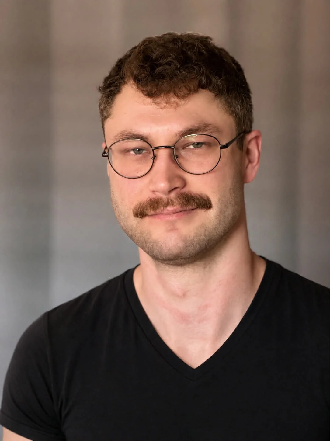 Portrait of a man with glasses and a mustache, wearing a black V-neck shirt, against a blurred neutral background.