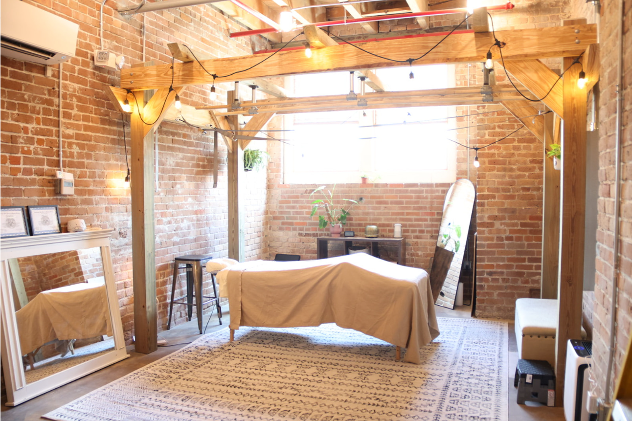 A massage therapy room with an exposed brick wall, string lights, a massage table with a white cover, a large standing mirror, and a small black cabinet with plants and decorative items.