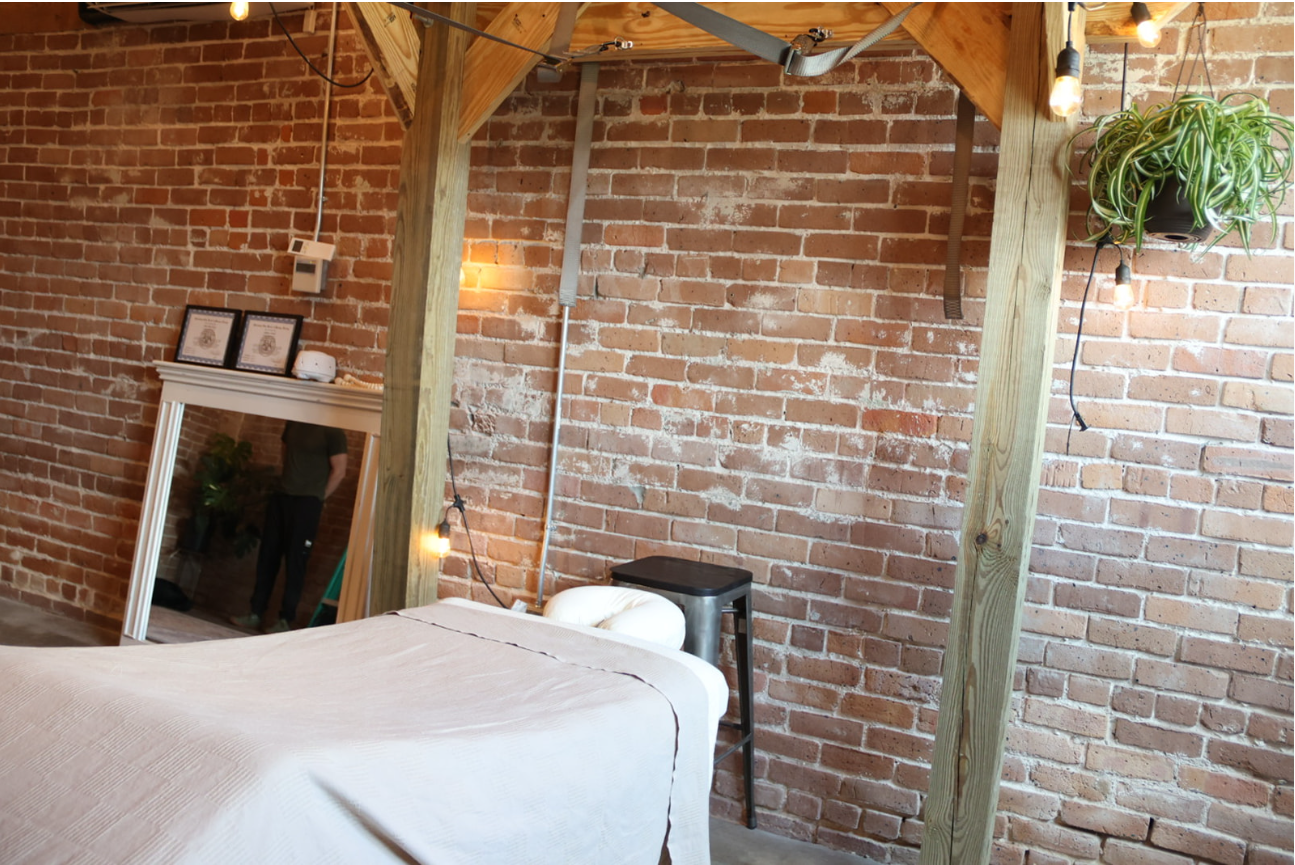 Interior of a room with exposed brick walls, wooden beams, a massage table with white linens, a black stool, a mirror leaning against the wall, and a potted plant hanging on the upper right corner. It is beautiful, rustic, and serene.
