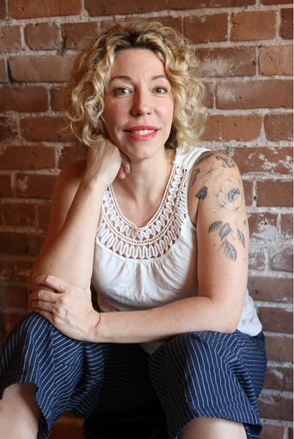 A woman with curly blonde hair, wearing a white sleeveless top with crochet detailing, sitting against a brick wall, smiling softly, with a tattoo of floral designs on her right upper arm.