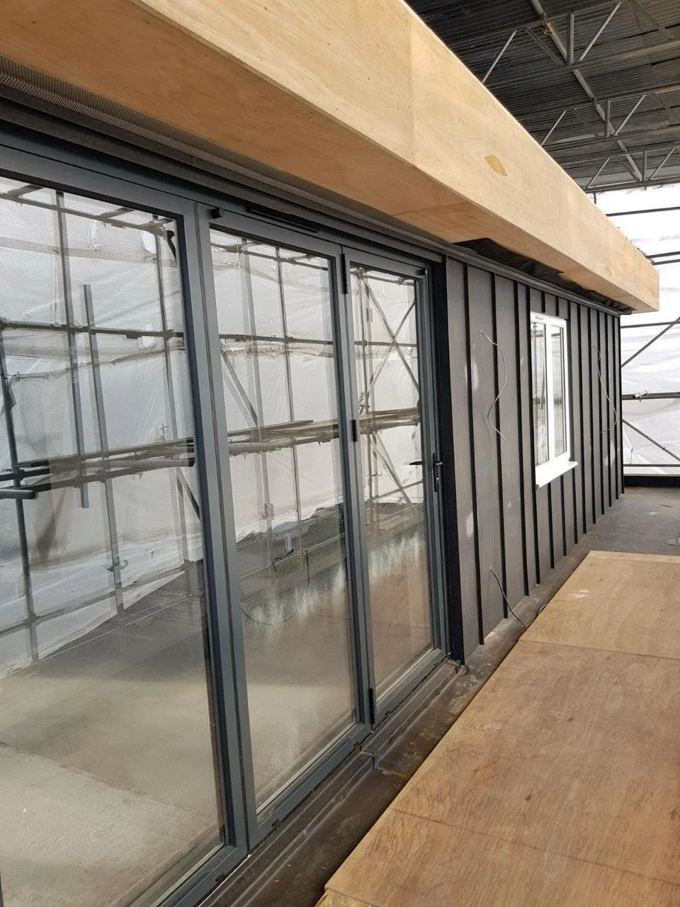 Construction site with a black exterior wall featuring sliding glass doors and a small white window, scaffolding reflected in the glass, and exposed ceiling structure.