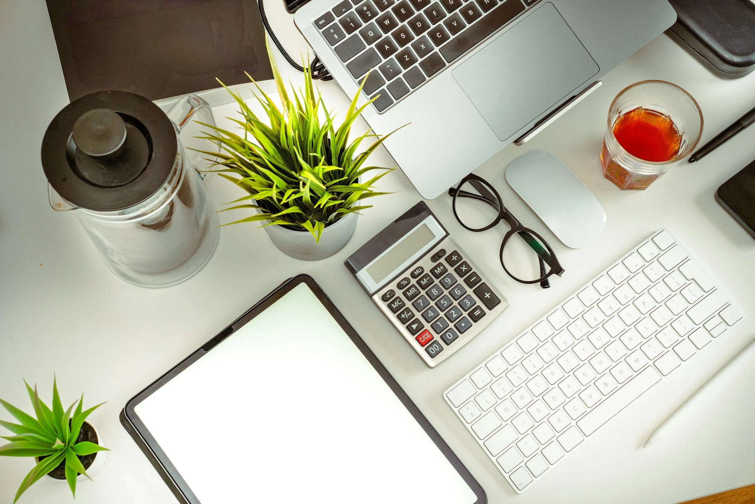 Computer workspace with a laptop, a white keyboard, a smartphone, a calculator, glasses, a pen, a glass of tea, a black case, a plant in a small pot, and a white mouse on a white desk.