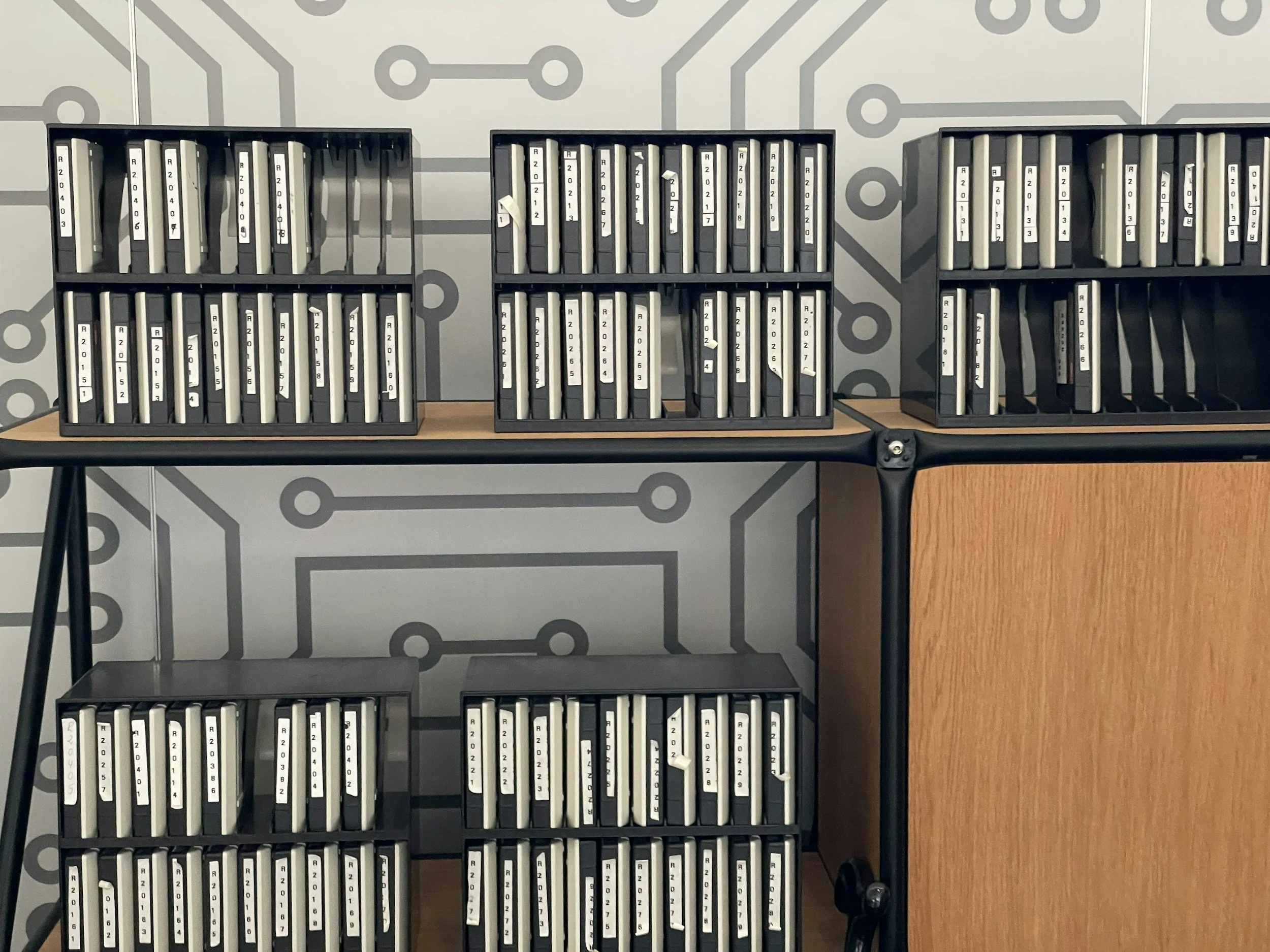 Three black storage trays filled with white index cards, labeled with numbers and letters, sitting on a black table against a wall with a circuit board pattern.