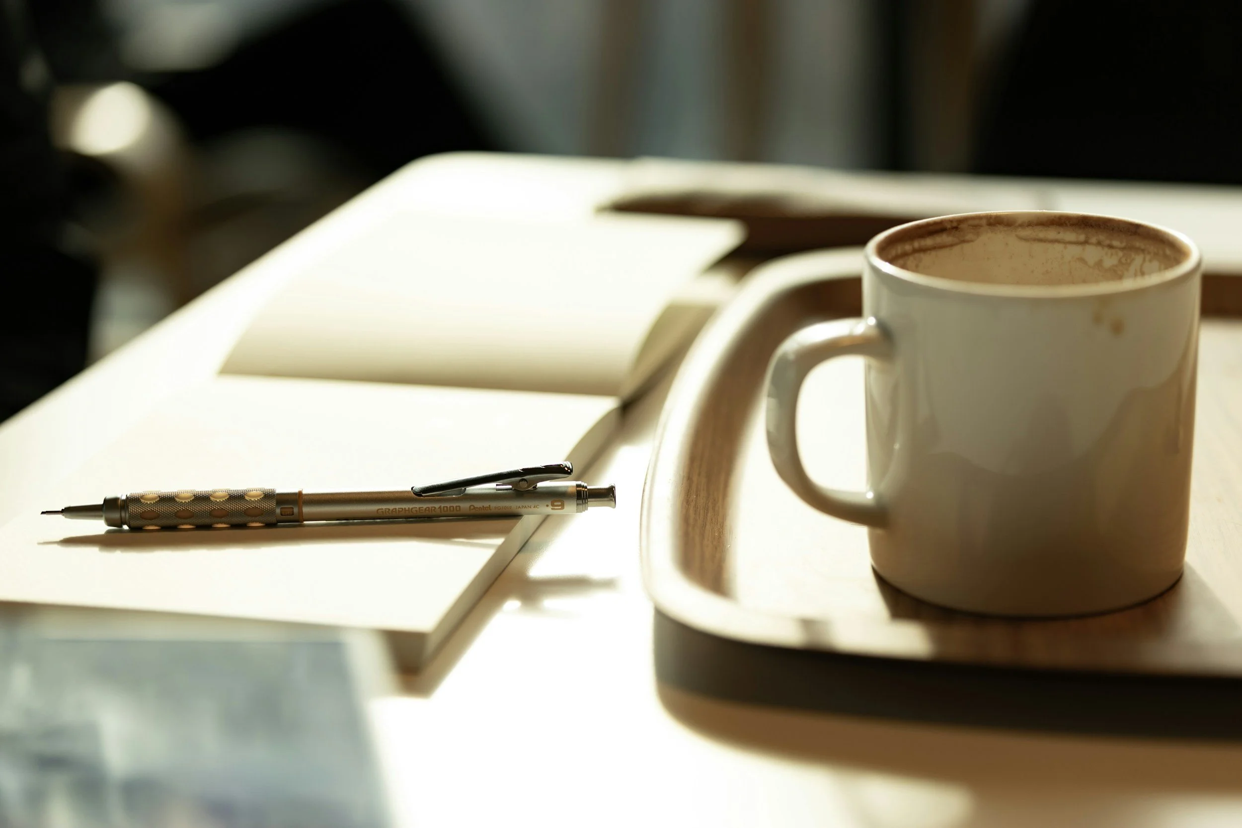 A white mug with coffee stains on the rim sitting on a desk with an open notebook and a pen in front of it.