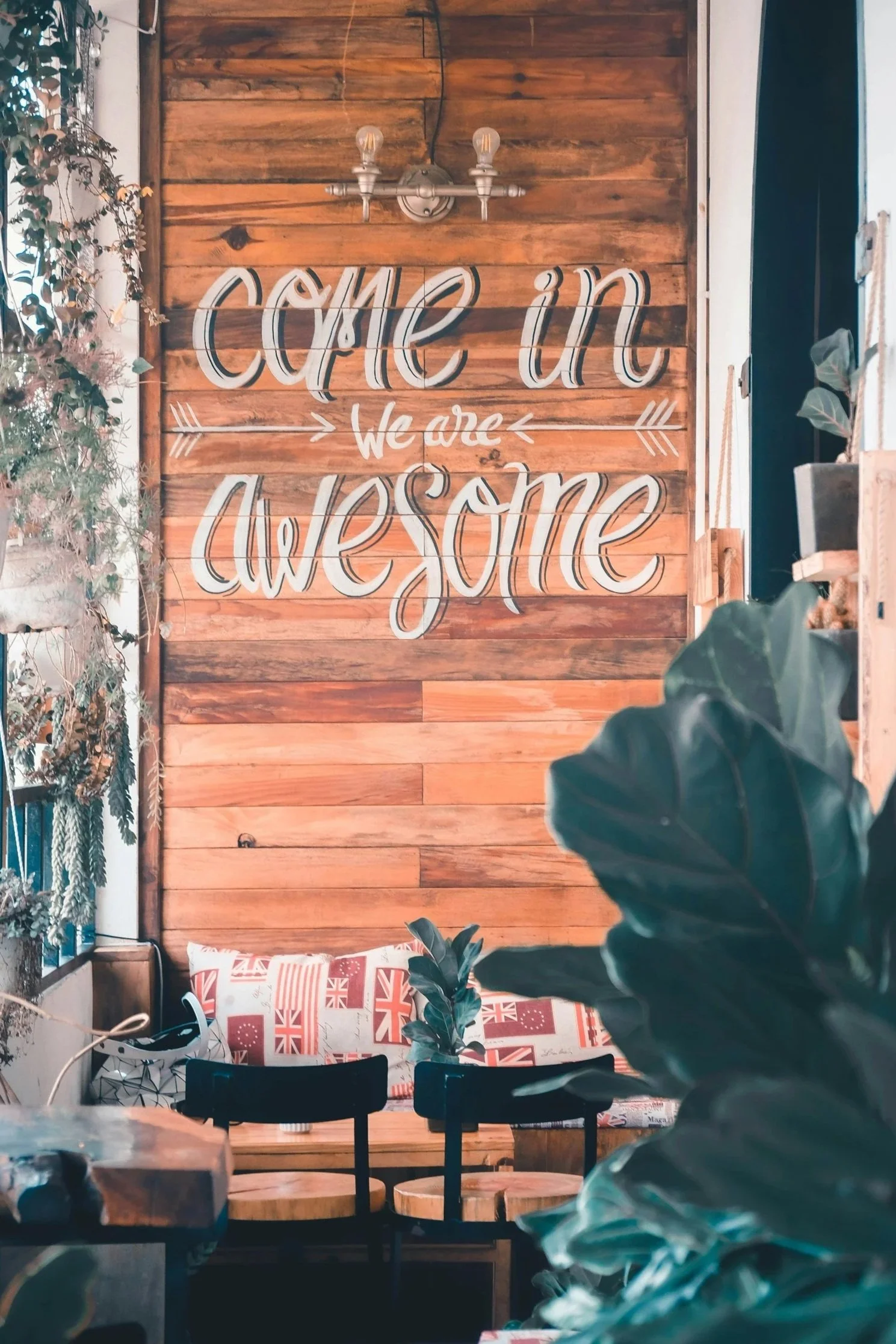 Interior of a cozy cafe or patio with a wooden wall, a sign that says 'Come in, We are Awesome,' surrounded by plants and outdoor furniture with British-themed cushions.