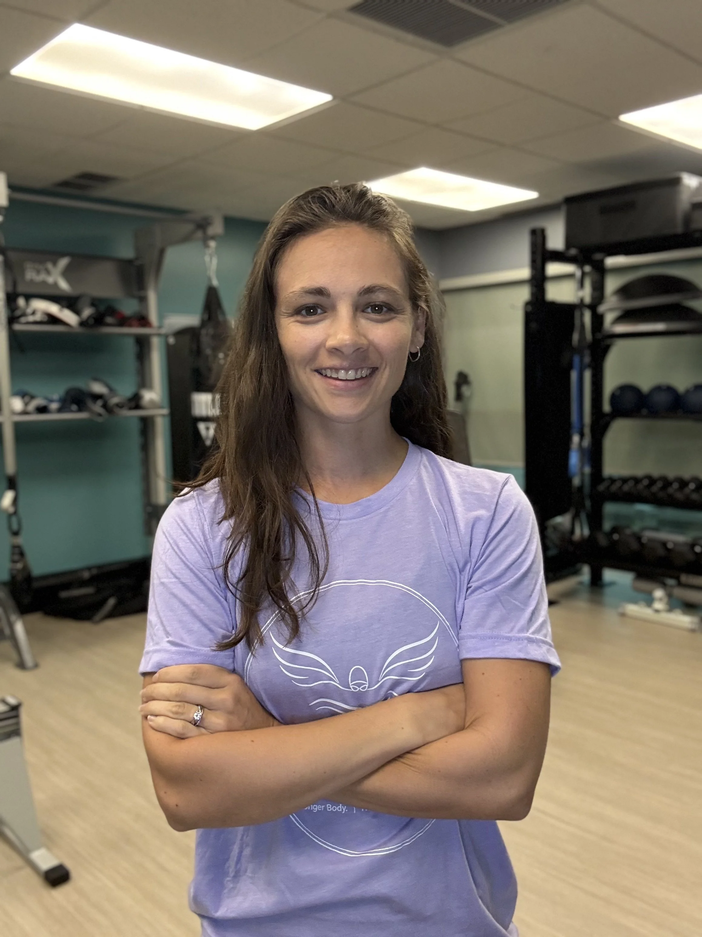 A smiling woman with long brown hair, wearing a purple t-shirt, arms crossed, standing in a gym with workout equipment in the background.