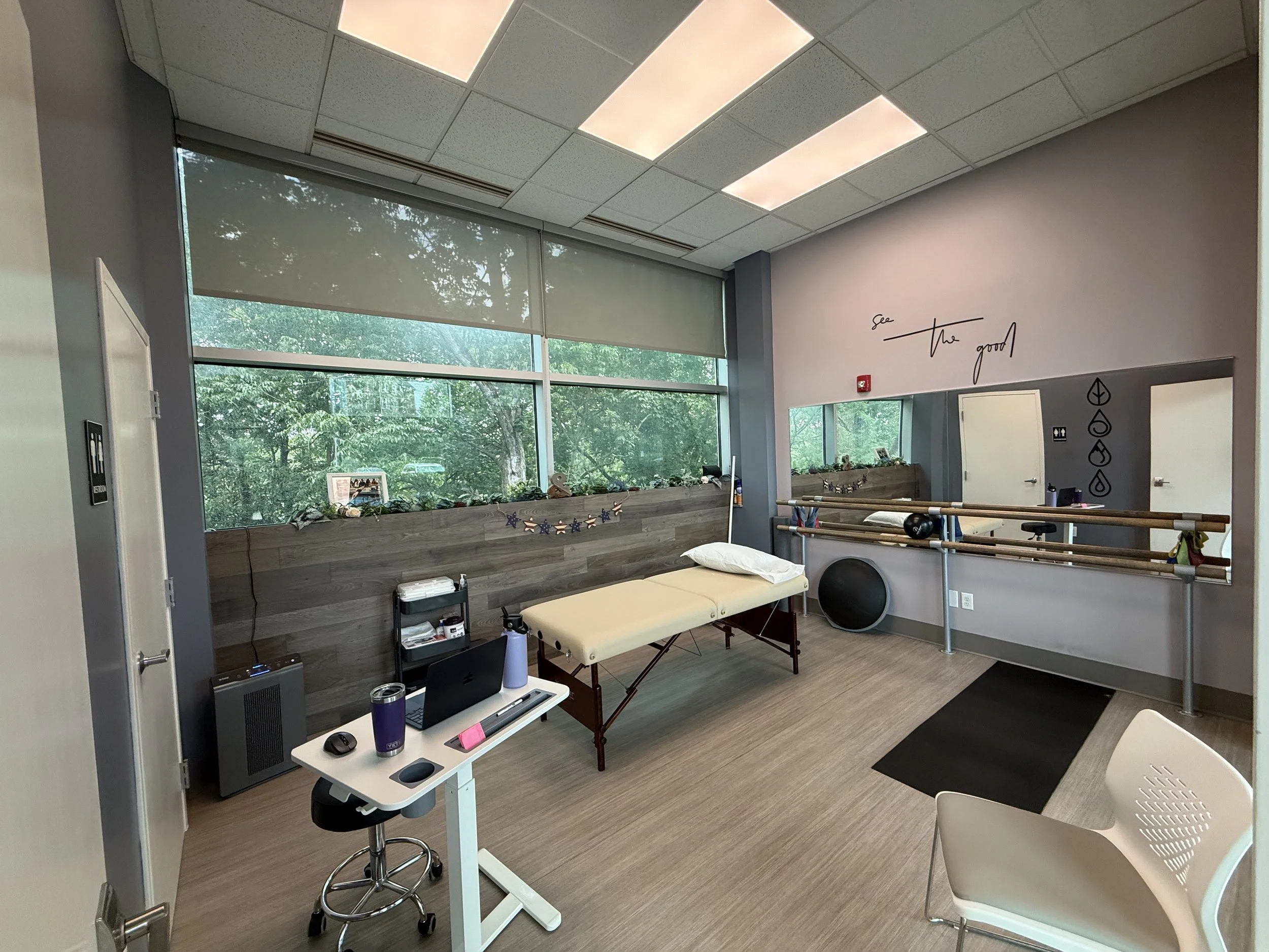 Interior view of a physical therapy or rehabilitation room with large windows showing greenery outside. The room has a treatment table with a pillow, a small table with a laptop, water bottle, and other items, and a chair. There is ballet barre attached to the wall, with the phrase 'see the good' written above, and some decorations and symbols on the wall.