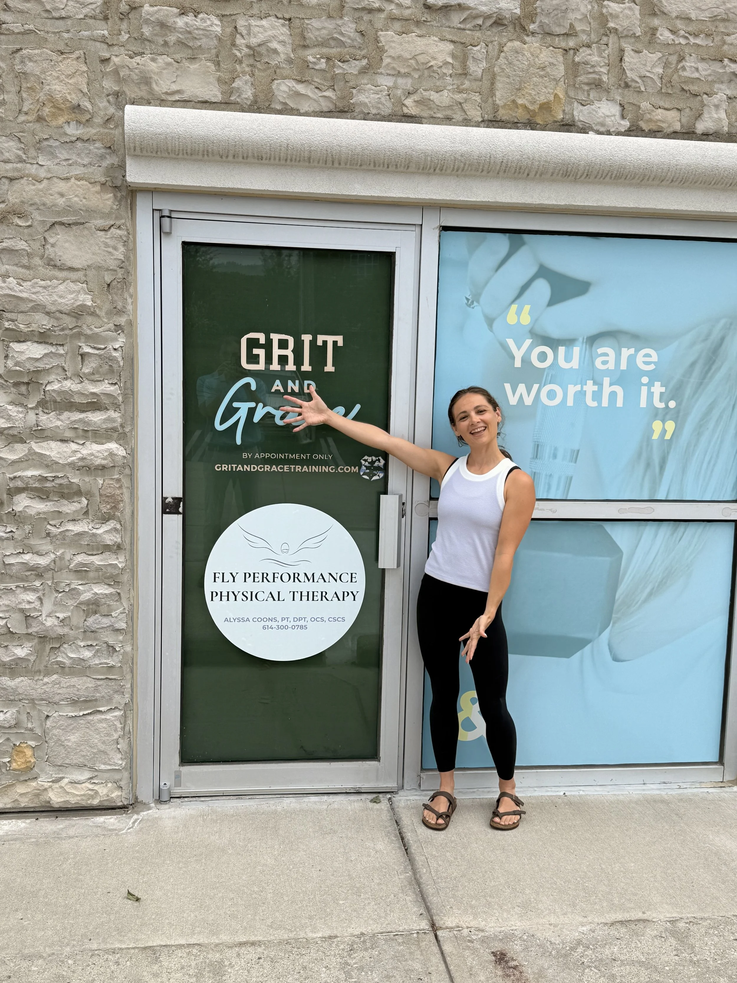 A smiling woman standing outside a building, pointing at a sign for Fly Performance Physical Therapy, with motivational text on the window that says  "You are worth it."