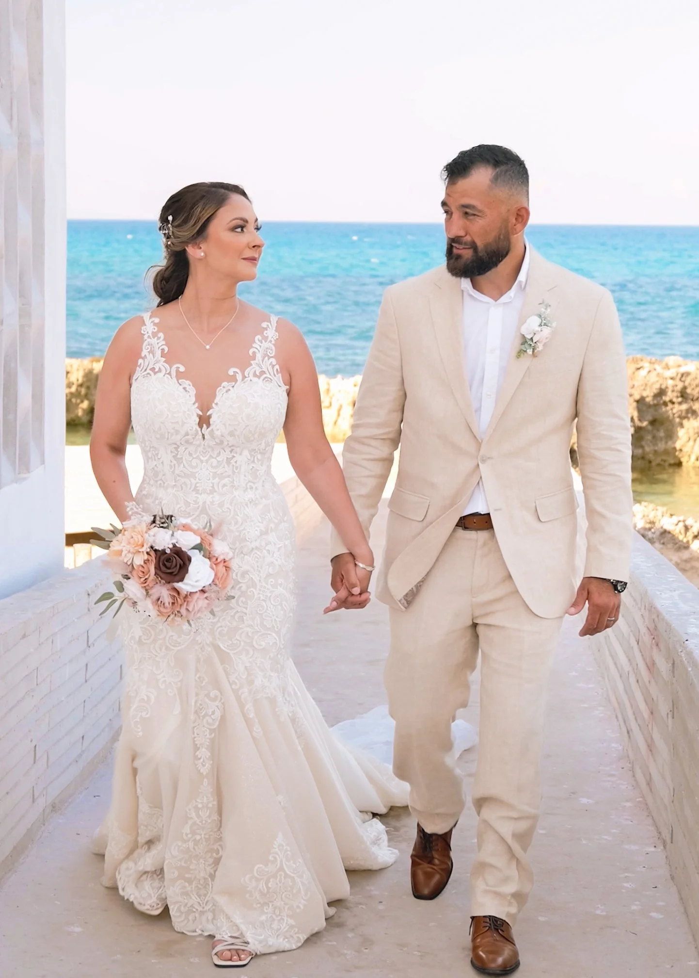 A bride and groom holding hands and walking on a white brick pathway with the ocean in the background, dressed in wedding attire.