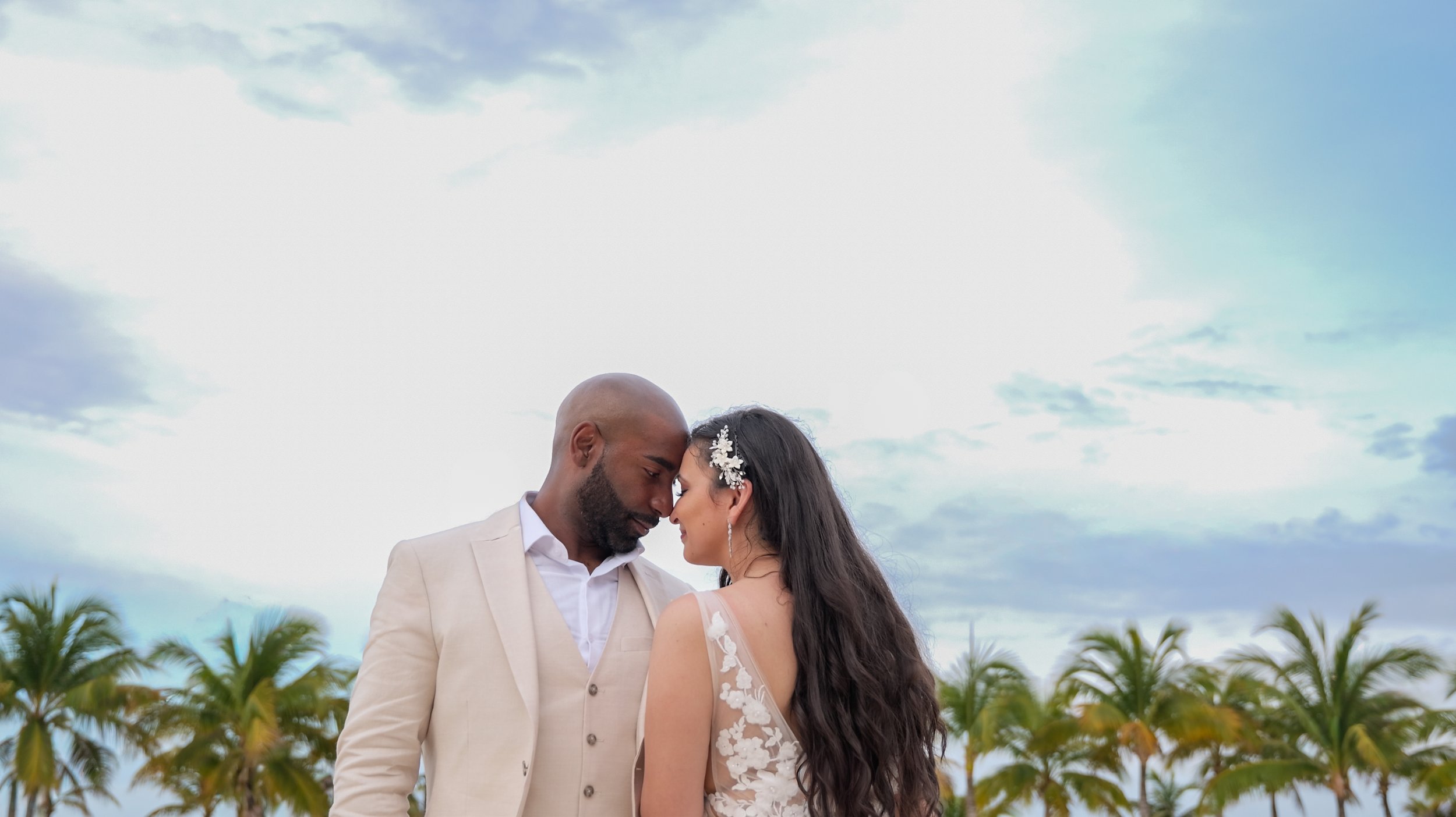 A loving couple, a man and a woman, standing close together outdoors with palm trees and a cloudy sky in the background, touching foreheads and gazing into each other's eyes.