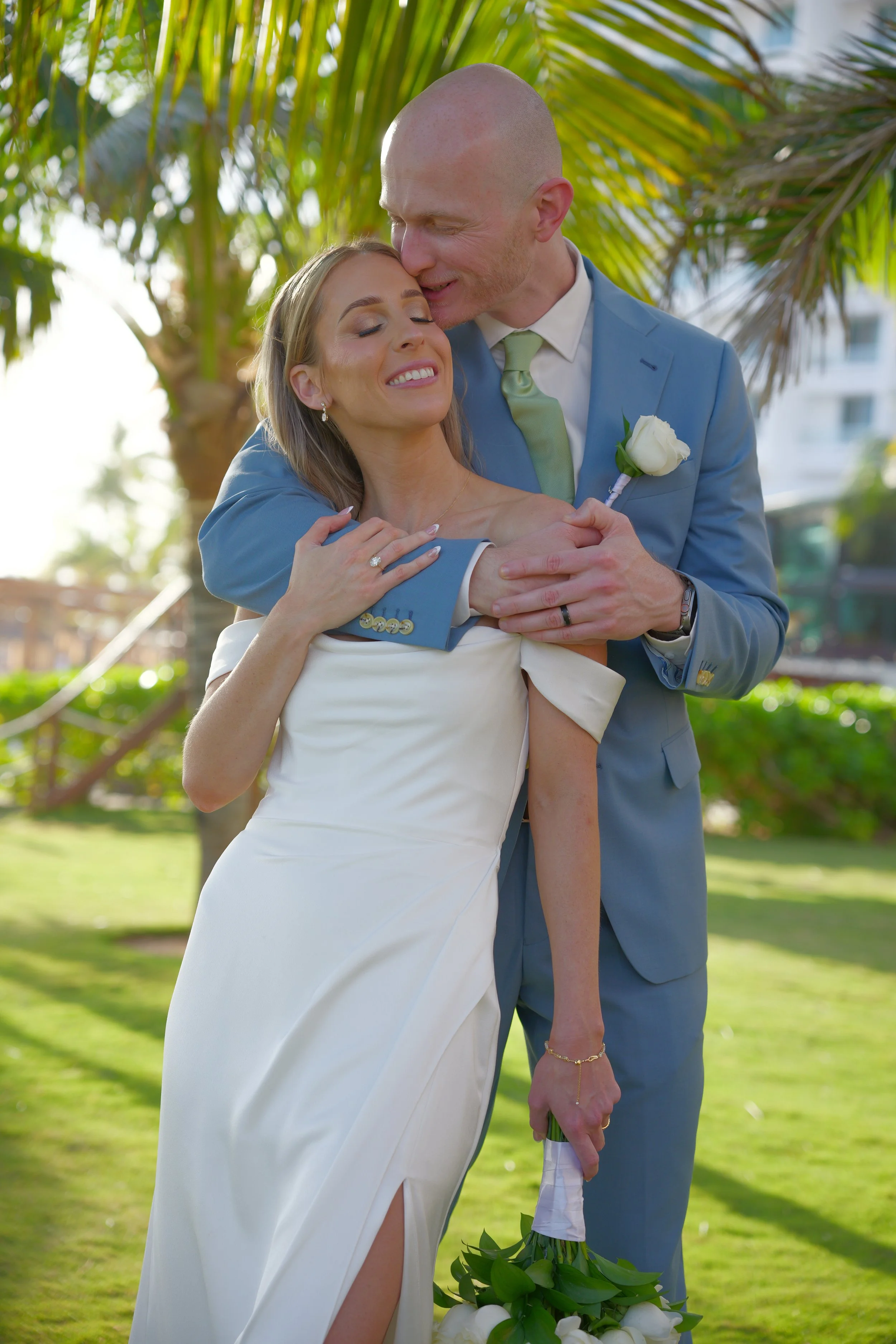 A bride and groom are embracing outdoors on a sunny day, with palm trees in the background. The groom is kissing the bride on her forehead, and she has her eyes closed with a smile. The groom is dressed in a light blue suit with a boutonniere, and th