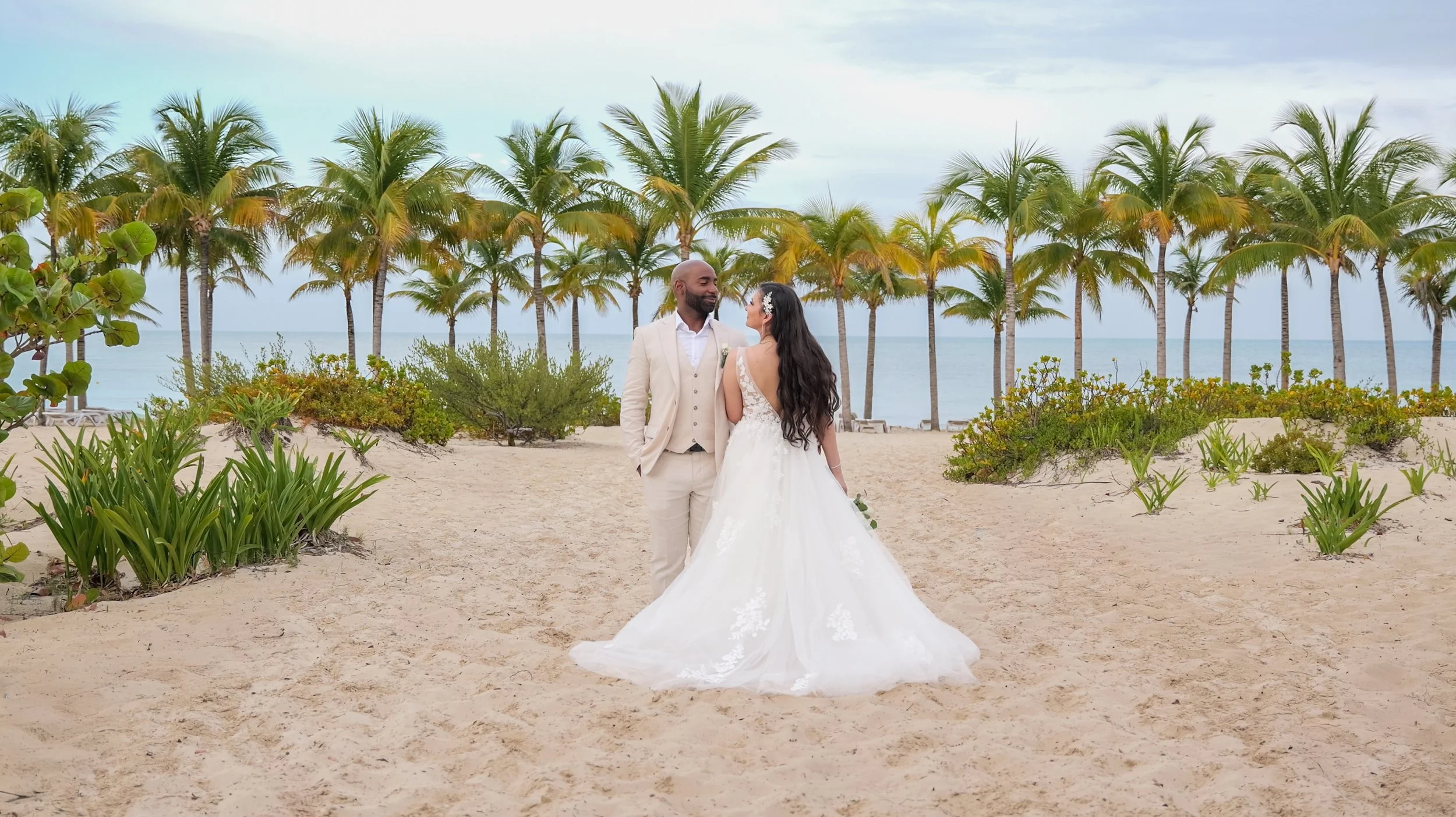A newlywed couple in wedding attire standing on a sandy beach with palm trees and the ocean in the background.