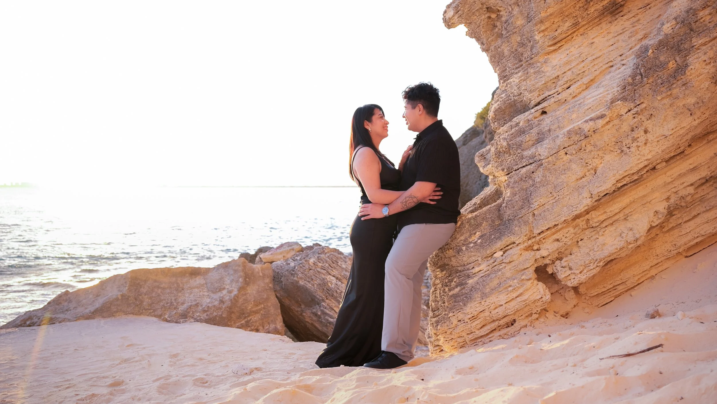 A couple stands close on a beach near large rocks, with an ocean and setting sun in the background, smiling and looking at each other.