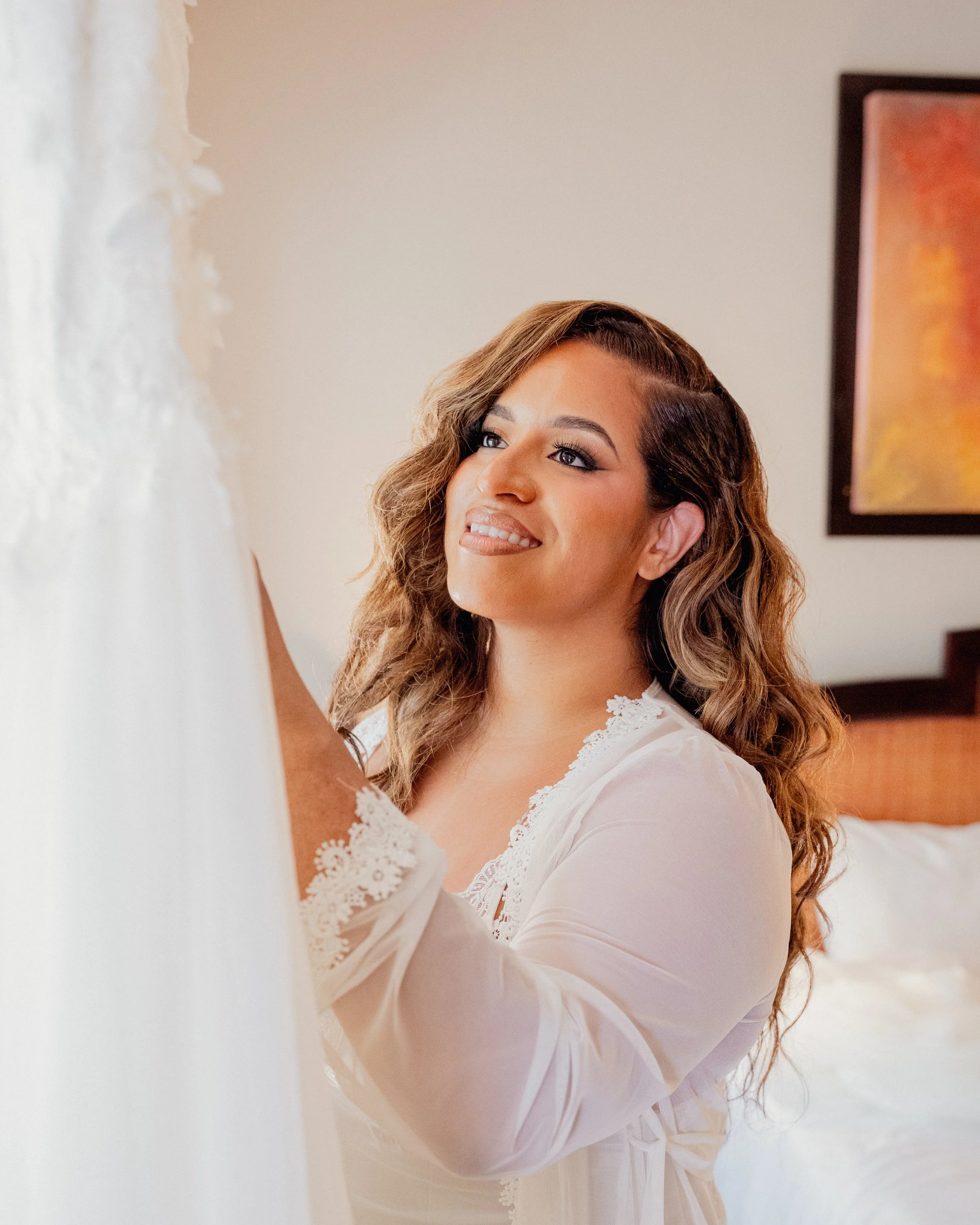 Young woman with curly brown hair and light skin wearing a white lace nightgown, standing in a bedroom, smiling and looking at herself in the mirror.