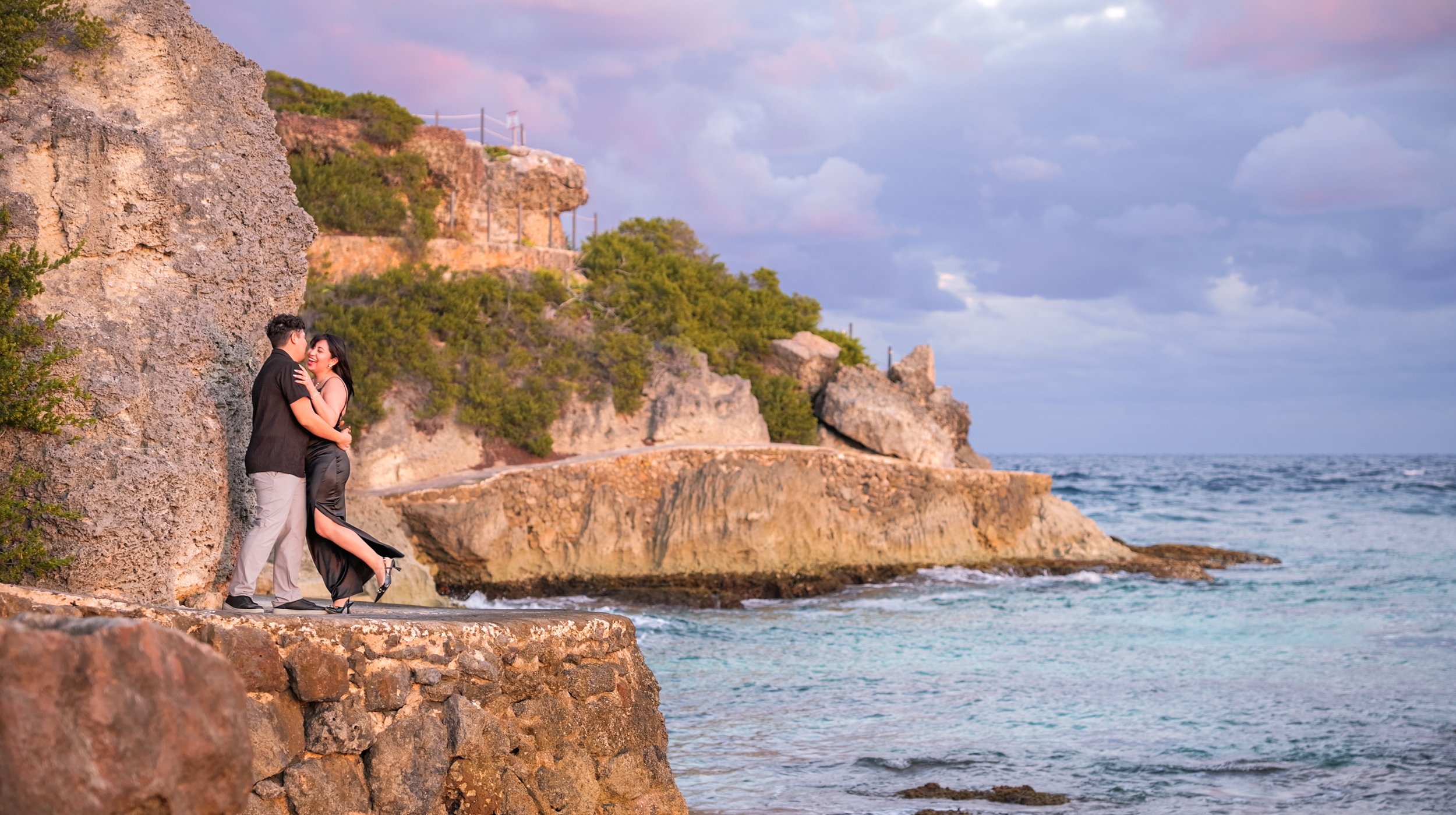 A couple is standing on a rocky coastal area, gazing at each other and smiling. The woman is wearing a black dress and high heels while the man is dressed in light-colored pants and a dark shirt. The background features cliffs with greenery, a cloudy
