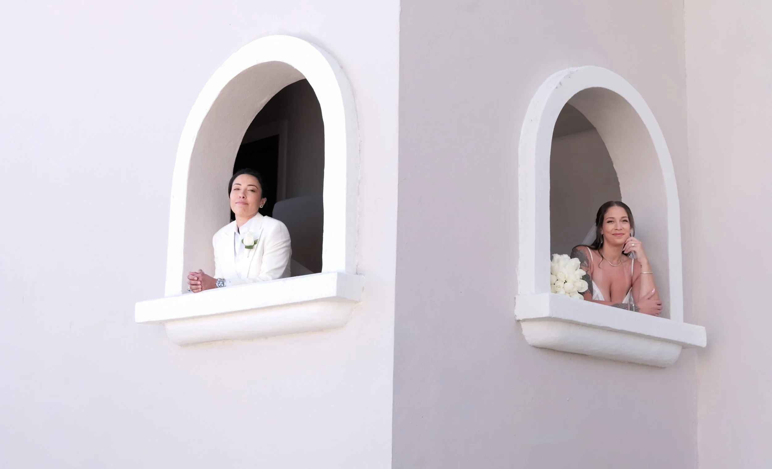 Two women leaning on white-walled arches, one is dressed in white and the other in a swimsuit, holding a bouquet of white flowers.