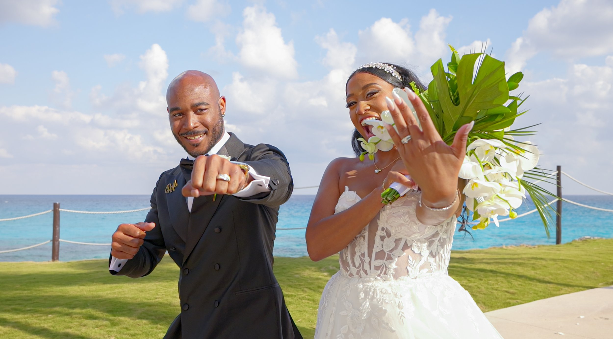 A bride and groom are celebrating on a beach, dressed in wedding attire, with the ocean and blue sky in the background.