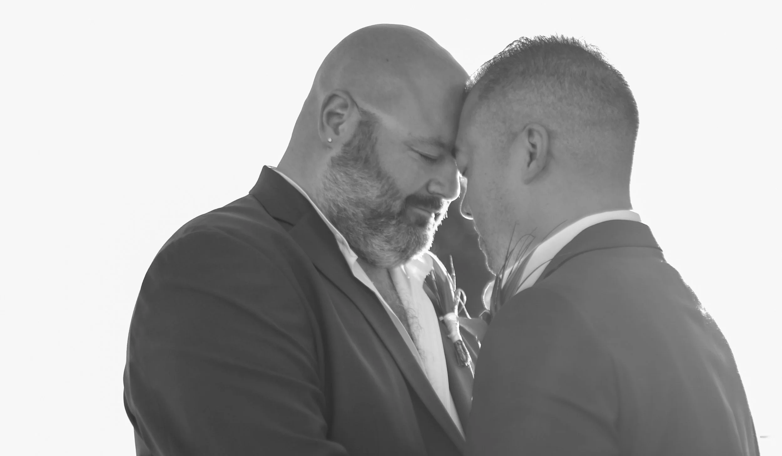 Two men in suits and ties share a tender moment, touching foreheads with closed eyes, against a plain background.