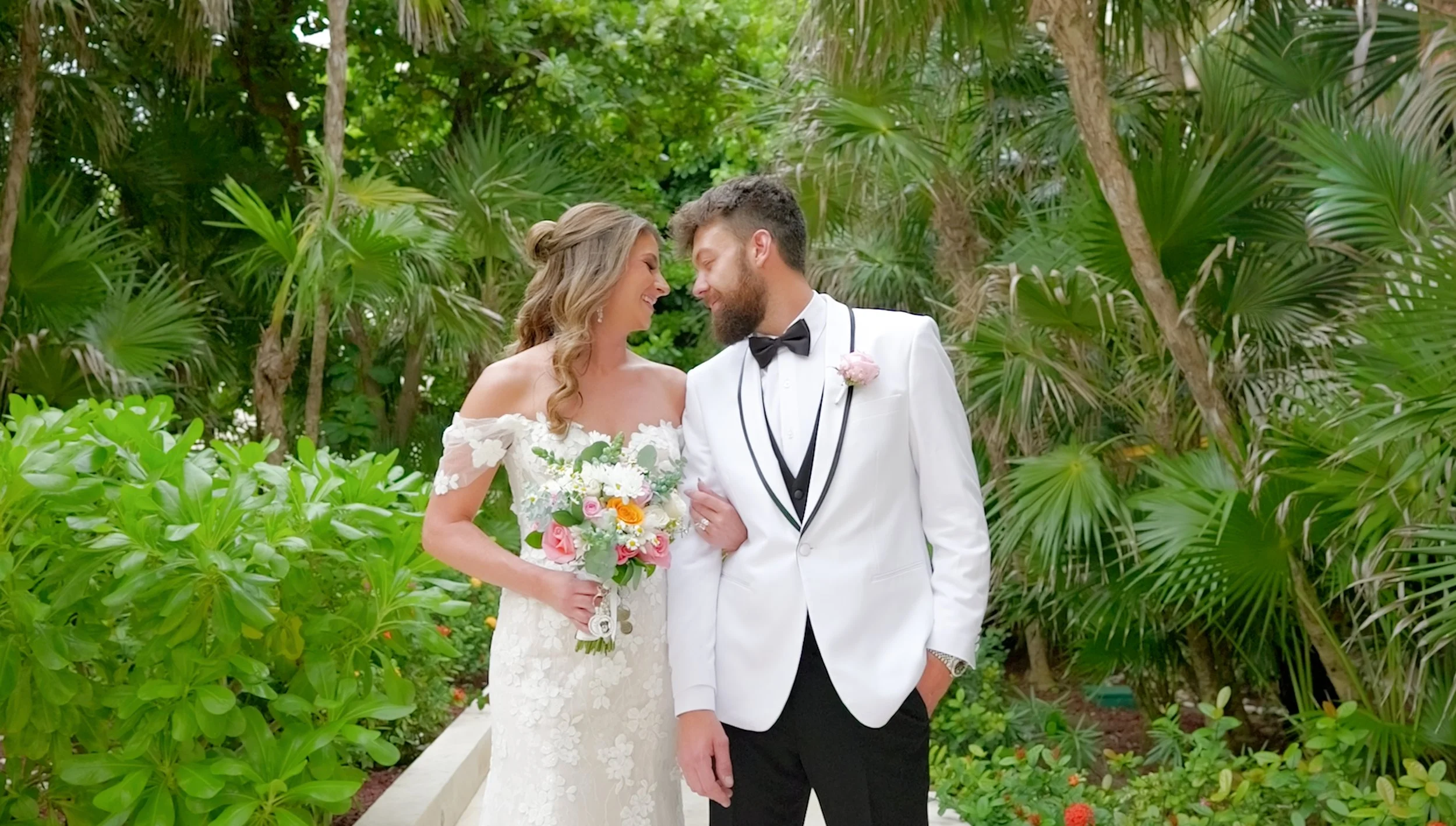 A bride and groom stand close together outdoors, smiling and looking at each other, surrounded by lush green tropical plants.