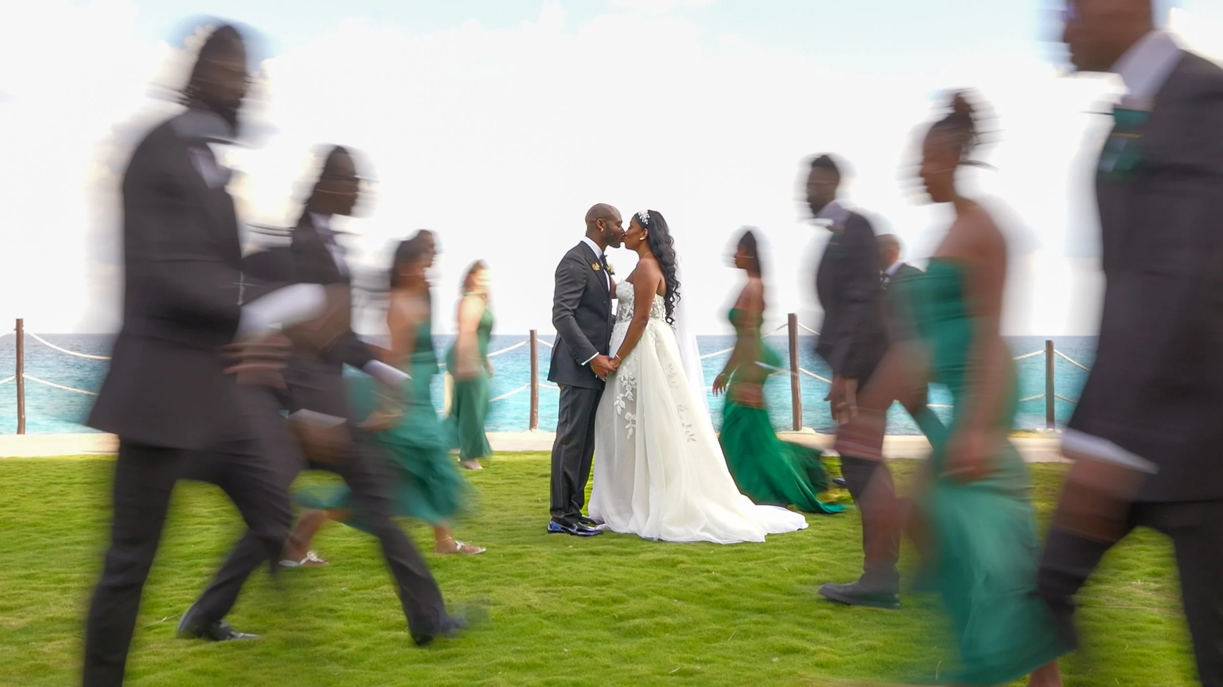 A wedding couple kissing under the sun, with guests walking past in colorful attire near a beach.