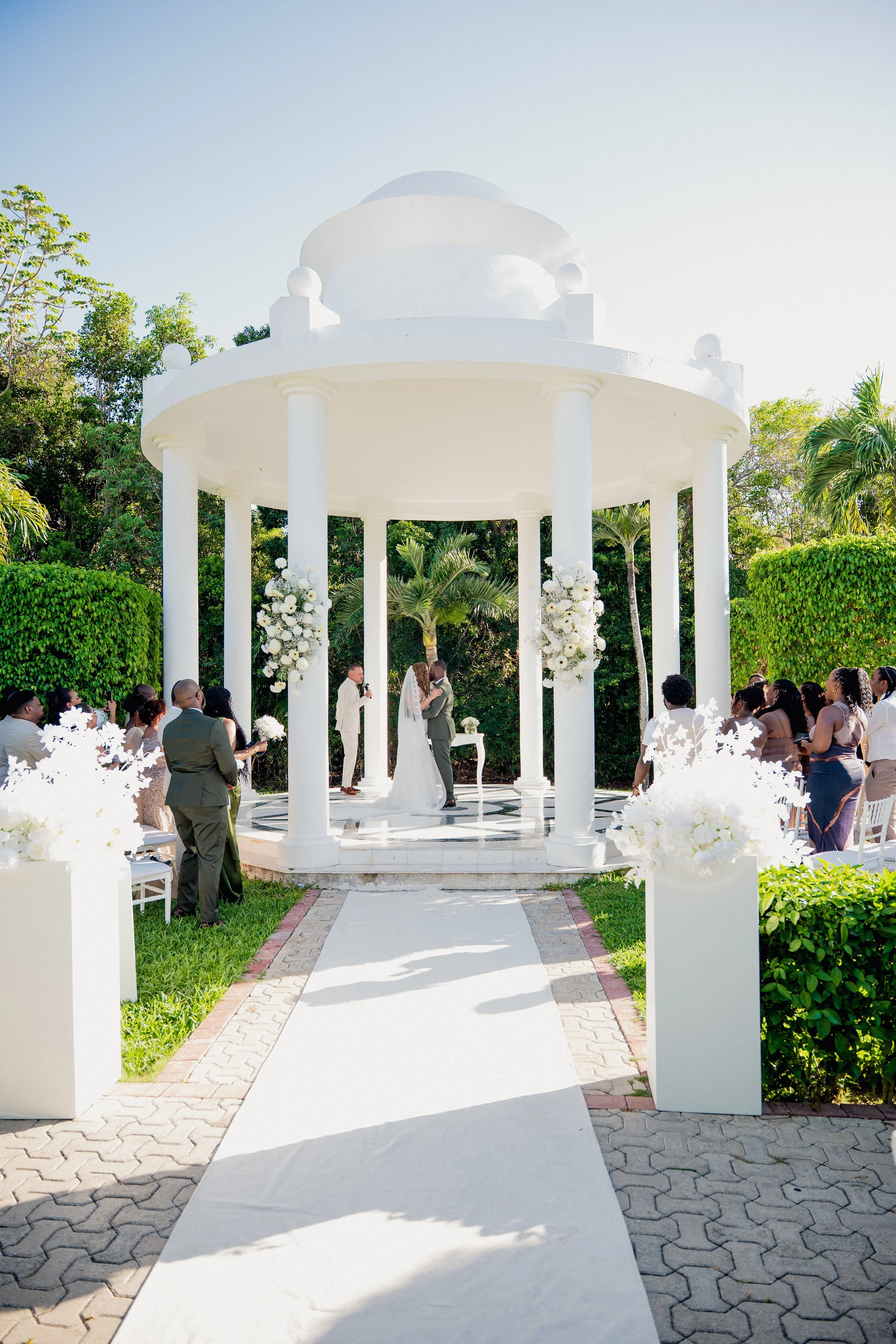 Couple getting married under a white gazebo, surrounded by guests, outdoor setting with trees and bushes, white aisle runner leading to the altar, and floral decorations.
