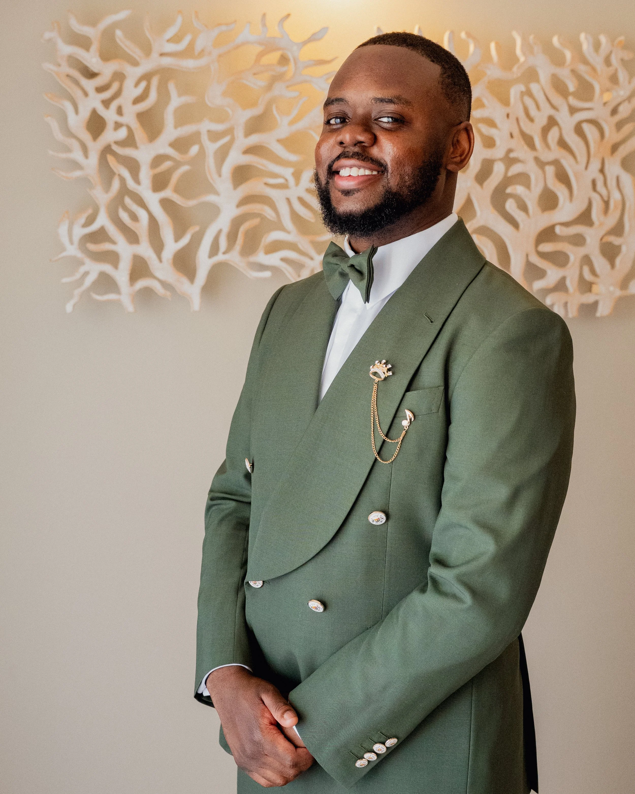 A man wearing a green tuxedo with a matching bow tie, standing in front of a decorative wall art with a light-colored background.