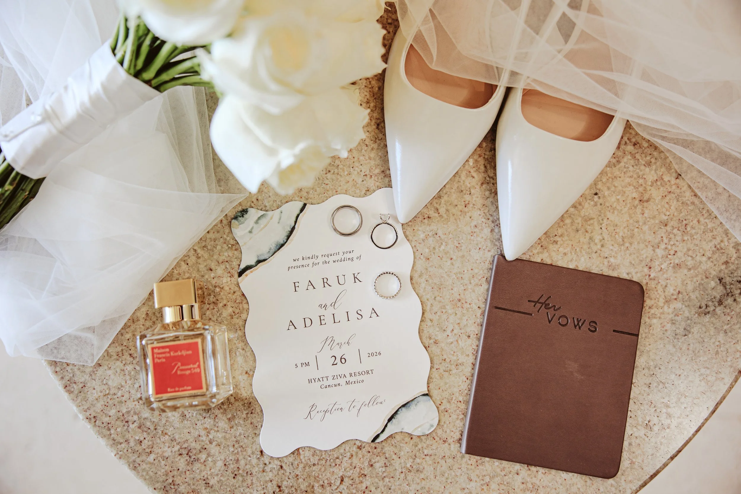 A wedding invitation card with the names Faruk and Adelisa, date March 26, 2026, at Hyatt Ziva Resort in Cancun, Mexico. Surrounding items include white high heels, a bouquet of white flowers, a small perfume bottle, wedding rings, a brown planner or