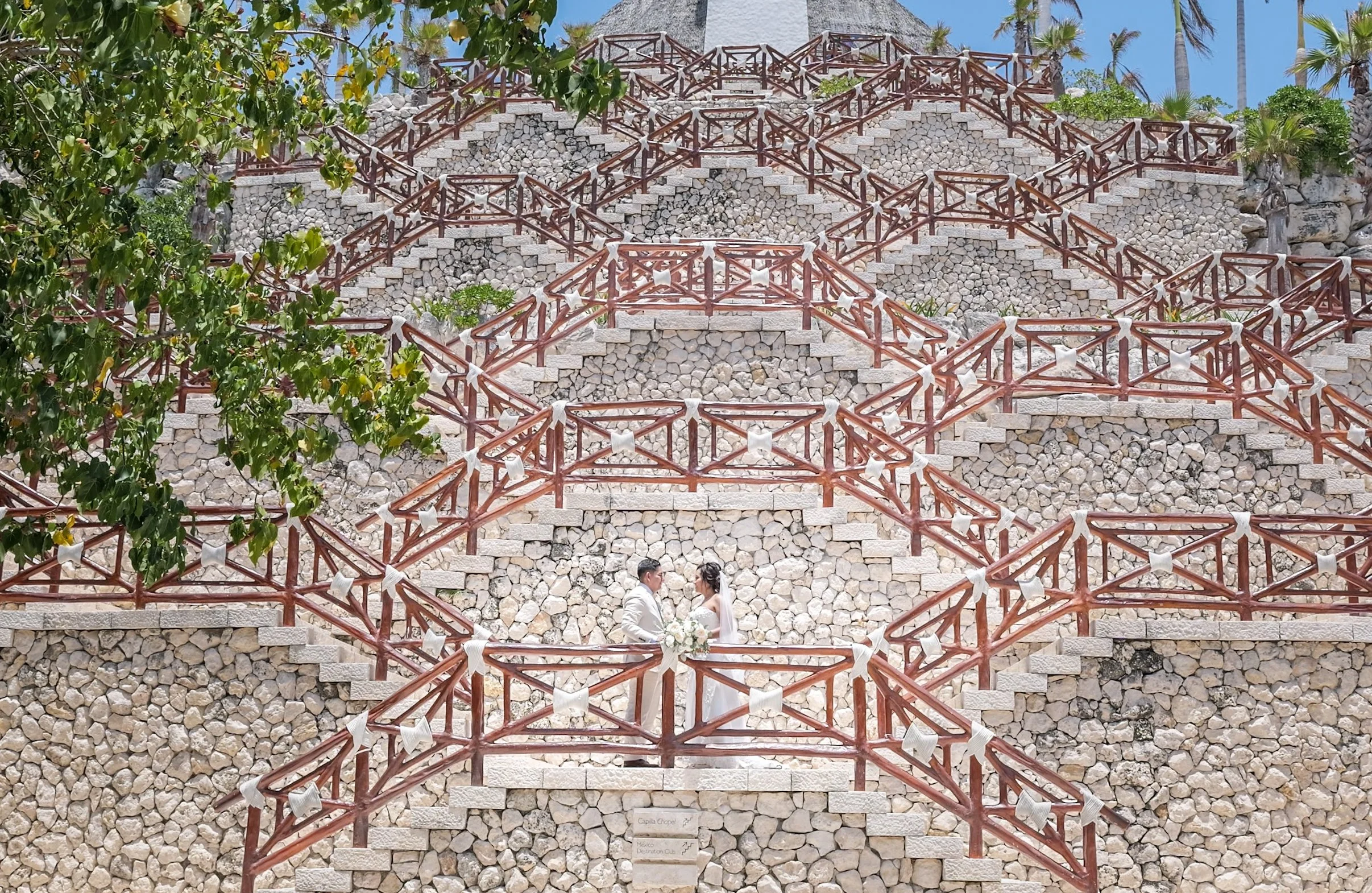 A couple dressed in wedding attire standing close together in front of a stone wall with a triangular pattern and wooden railings, surrounded by green trees and palm trees, likely at a wedding venue.