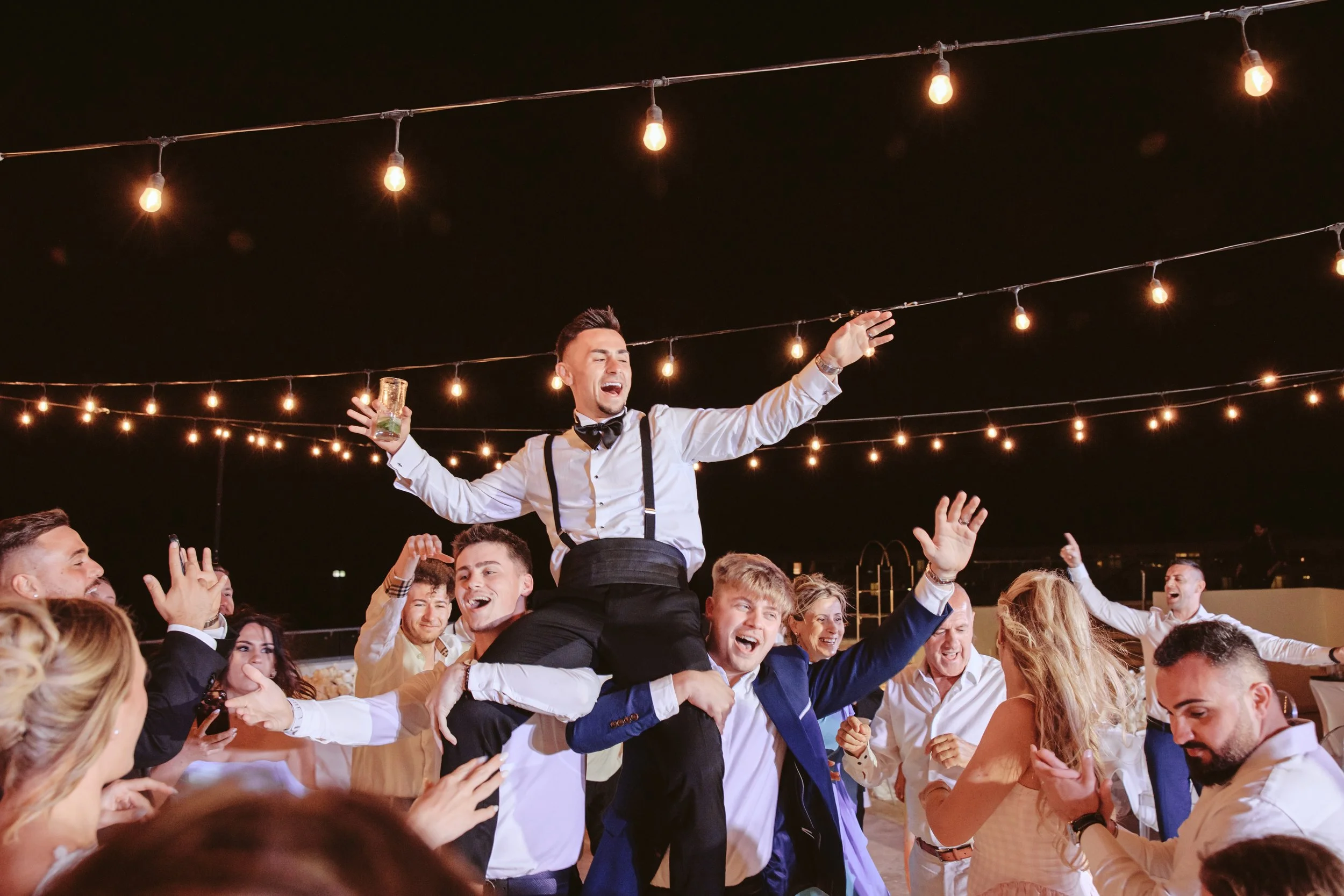 A group of people celebrating at a party outdoors at night, with a man wearing a tuxedo and suspenders sitting on a person's shoulders, holding a drink and dancing among smiling guests under string lights.