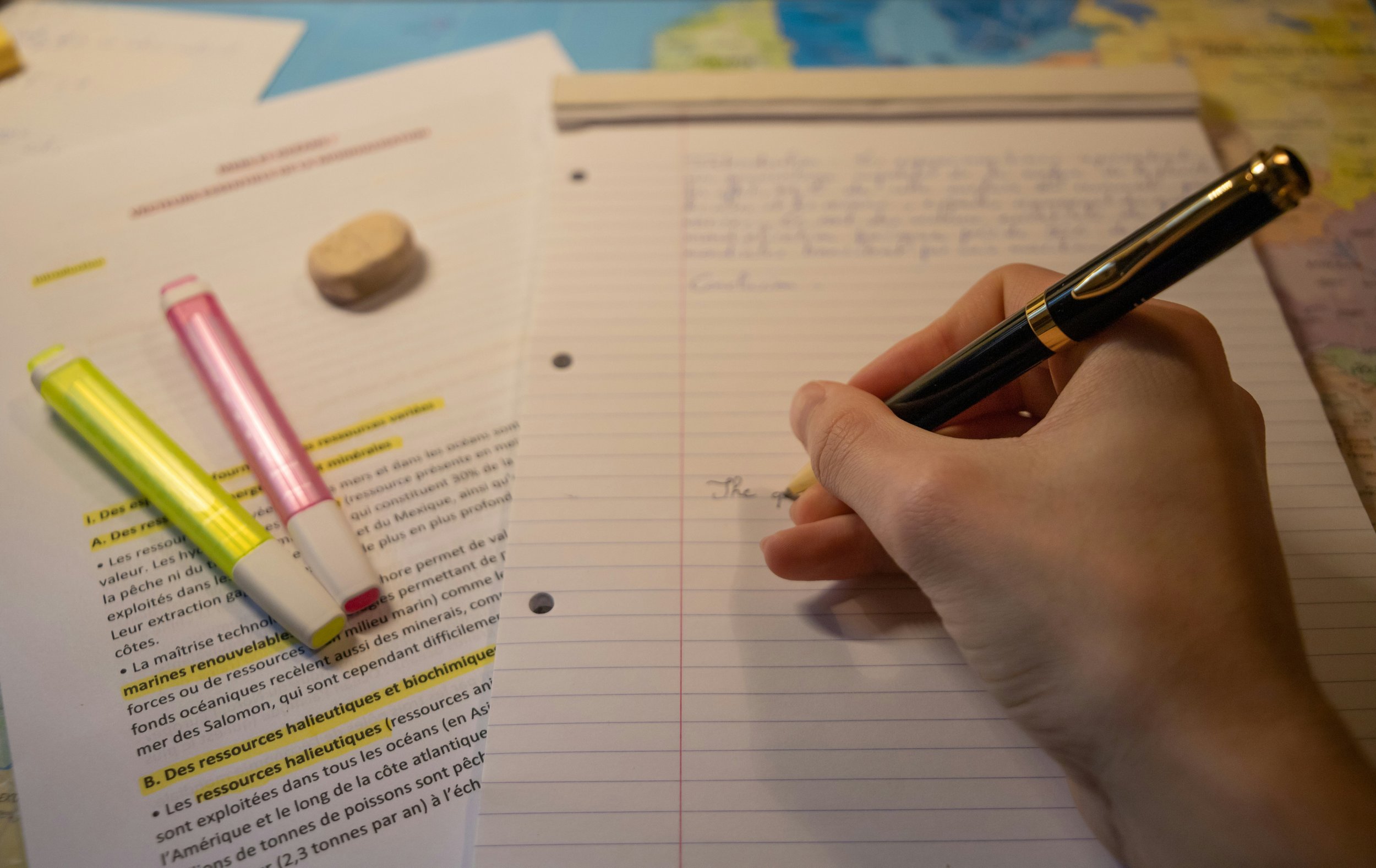 A person's hand writing on lined notebook paper with a gold and black pen, surrounded by highlighted texts and additional papers with typed and highlighted notes. There are markers and an eraser on the table.