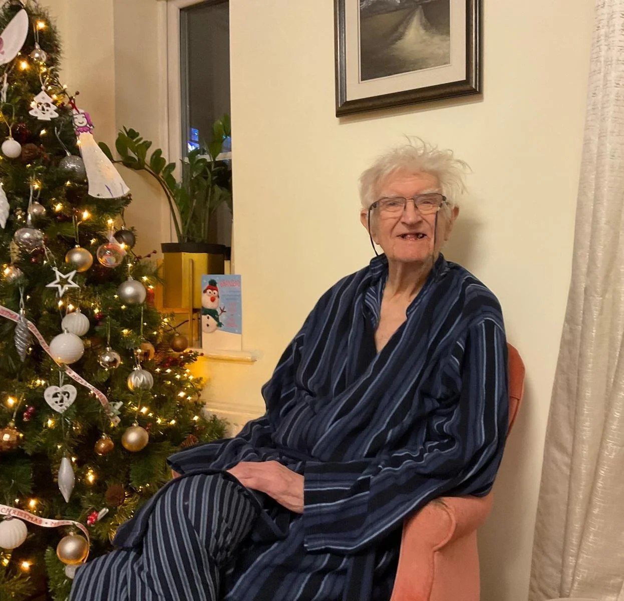 An elderly man with white hair, glasses, and missing teeth, sitting in a pink armchair beside a decorated Christmas tree with lights and ornaments.