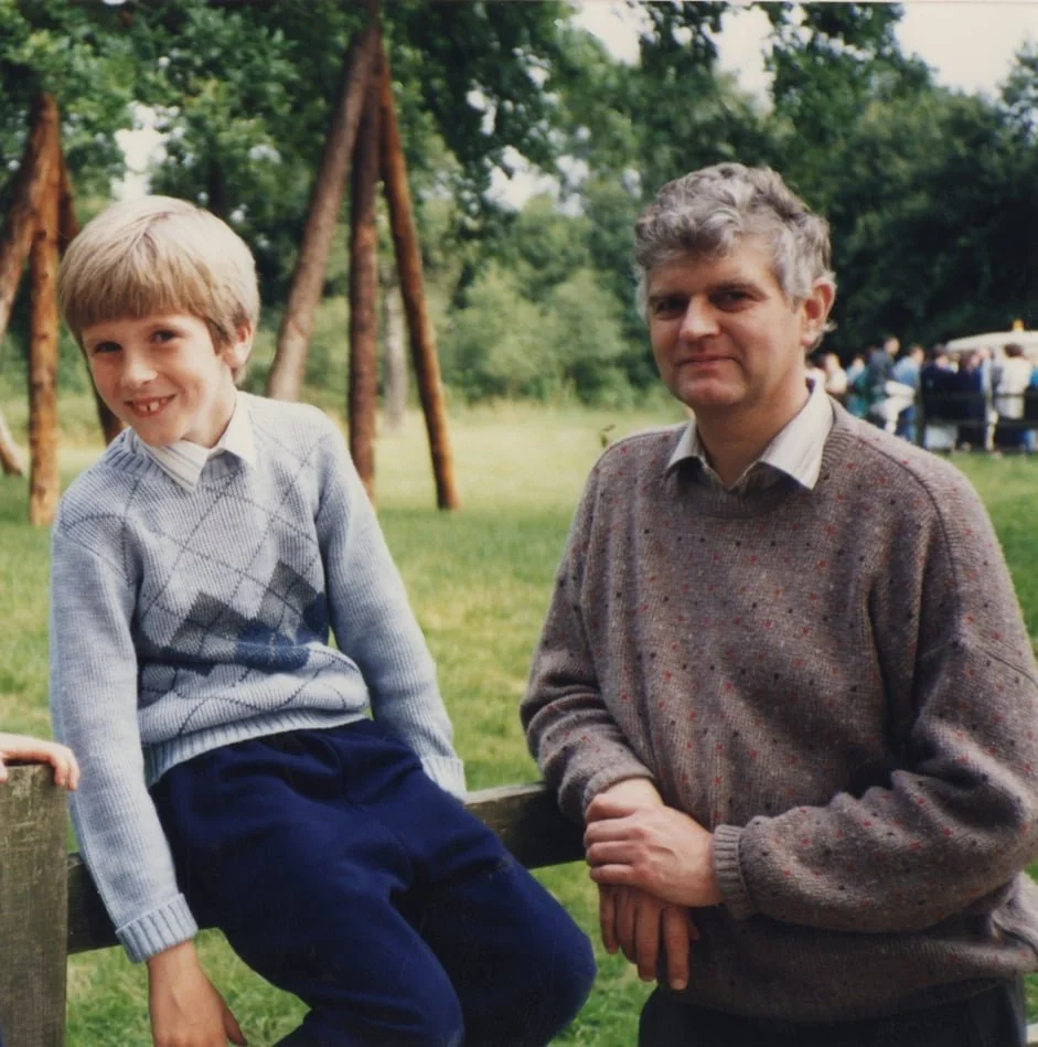 A young boy sitting on a wooden railing next to an adult man outdoors, with trees and a group of people in the background.