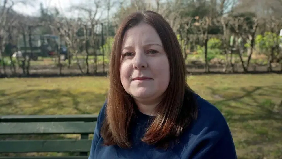 A woman with shoulder-length brown hair sitting on a park bench, with trees and a fence in the background, on a sunny day.