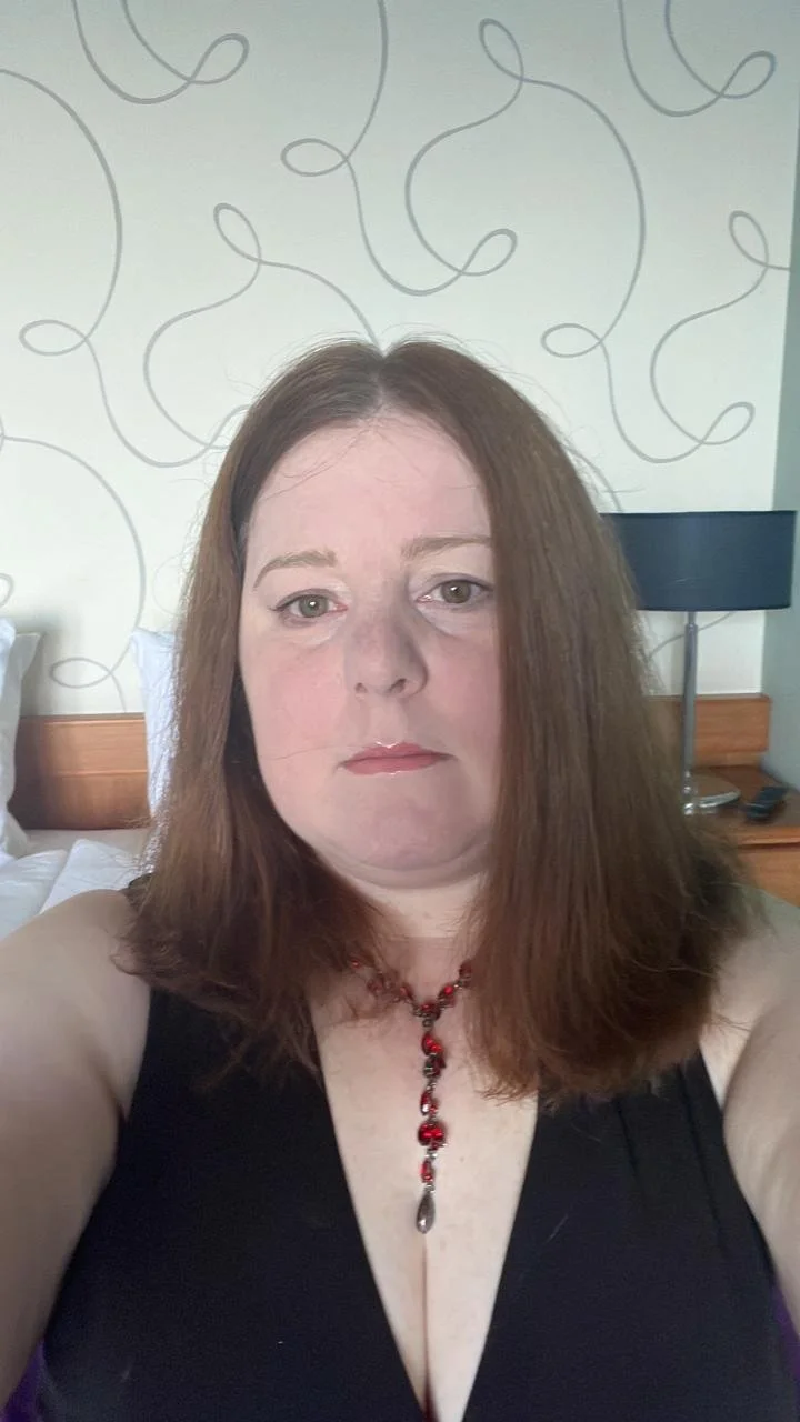 A woman with shoulder-length brown hair and light skin taking a selfie in a hotel room. She is wearing a black top with a deep neckline and a red necklace with a pendant. In the background, there is a bed with white sheets, a wooden headboard, a black table lamp, and wallpaper with a line design pattern.