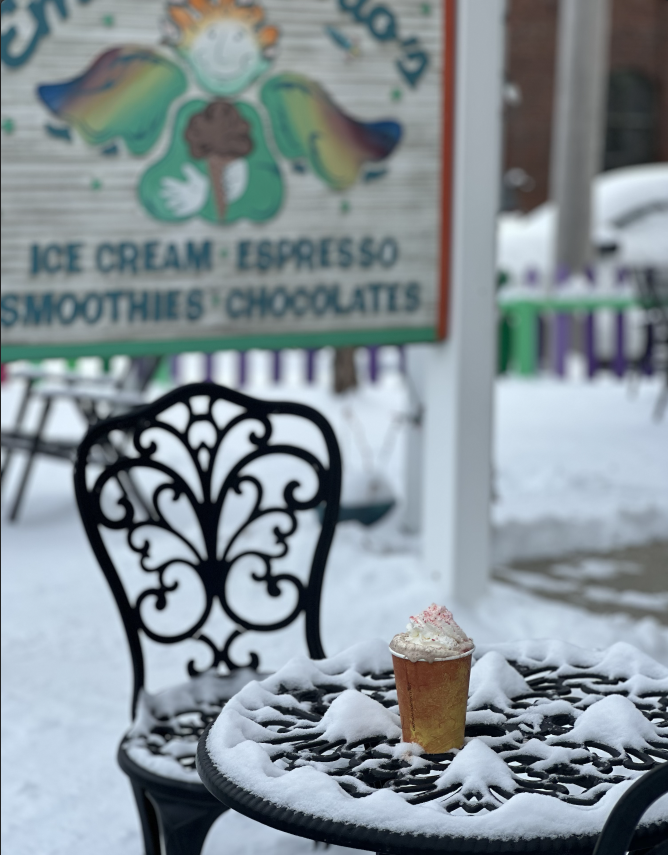 A drink in a cone-shaped cup with whipped cream on top sits on a snow-covered black metal outdoor table, with a decorative black metal chair behind it. In the background, snow covers the ground and a sign for a shop offering ice cream, smoothies, chocolates, and espresso is visible.