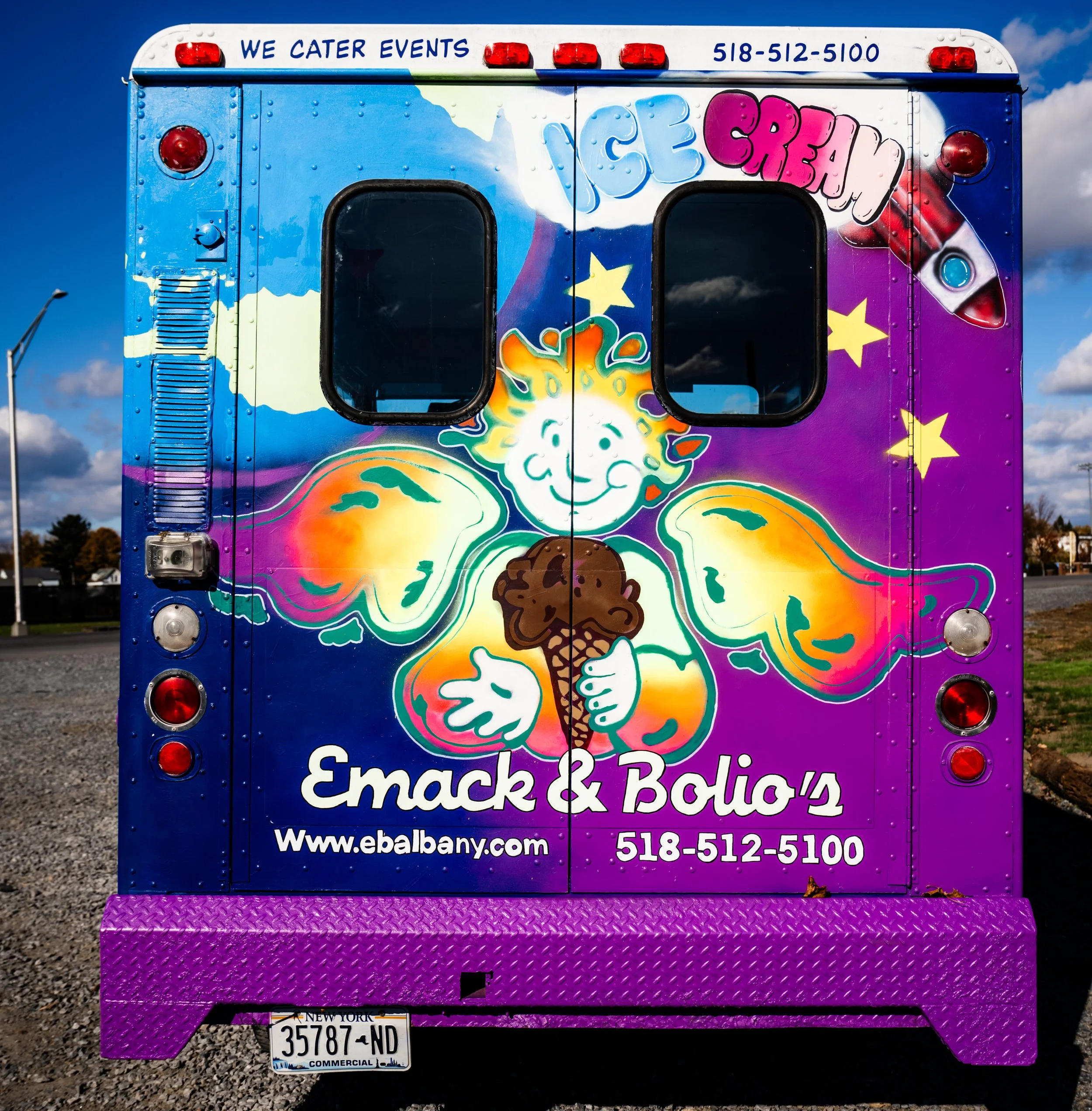 The back of a food truck with colorful graffiti art of a smiling ice cream cone and the words "Ice Cream" on the top. The truck has contact information and mentions catering events, along with a website and phone number.