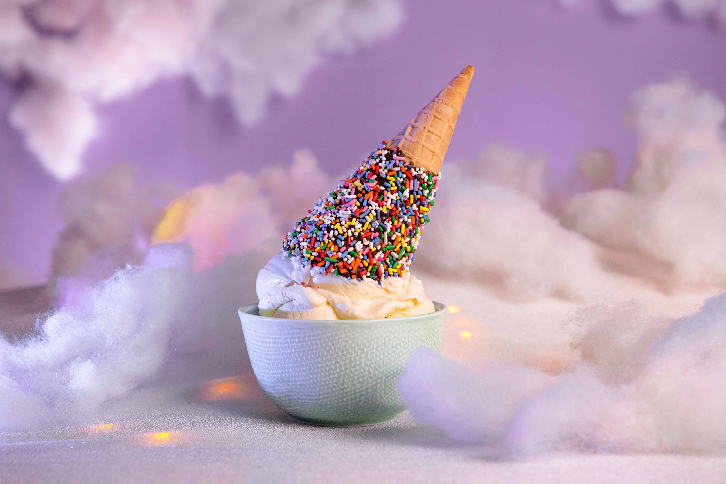 A cup of vanilla ice cream topped with a waffle cone and colorful sprinkles, set against a soft, fuzzy background with pastel colors.