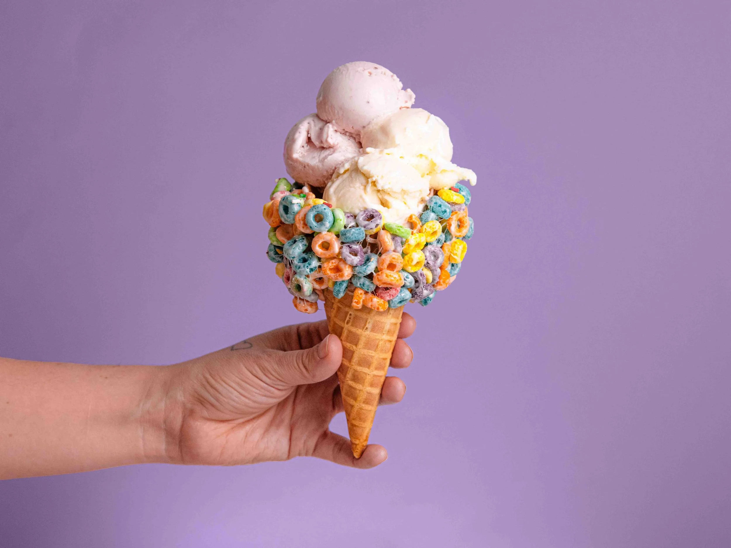 Hand holding an ice cream cone with three scoops of pink and white ice cream, decorated with colorful cereal loops against a purple background.