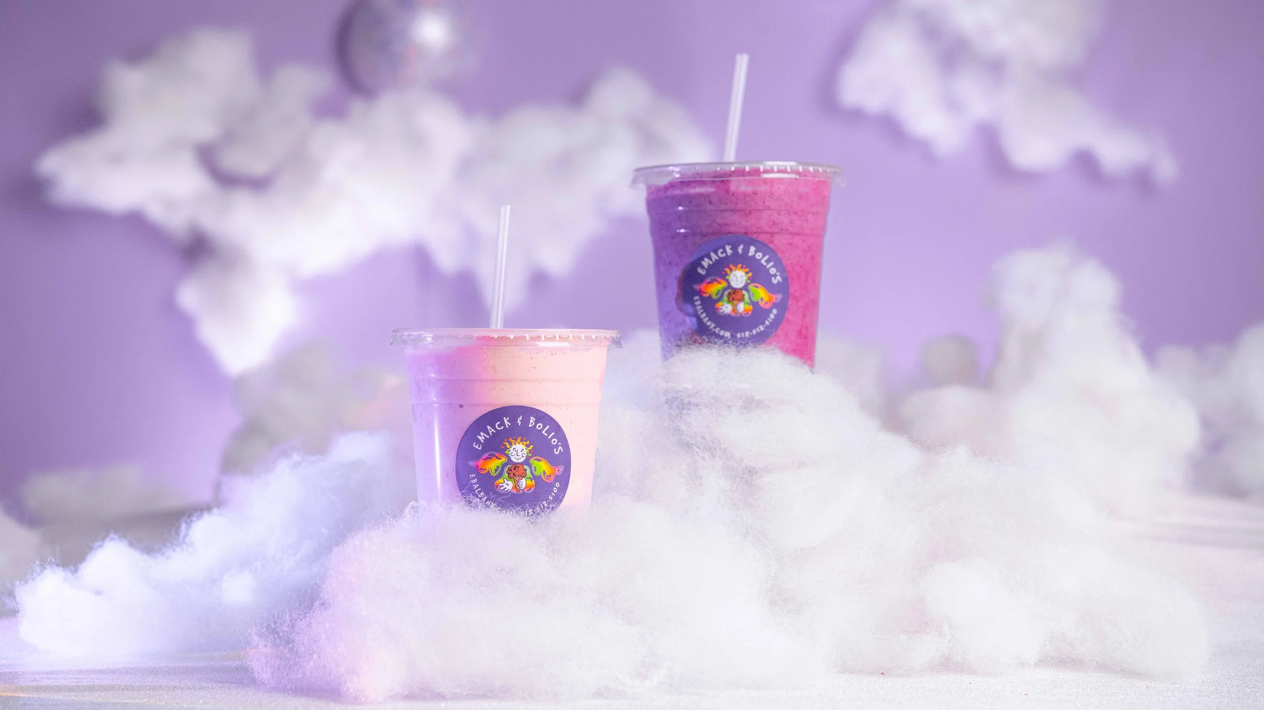 Two colorful smoothies, pink and purple, in clear cups with straws, placed on fluffy white clouds with purple background, featuring a logo with a cheerful character and rainbow wings.