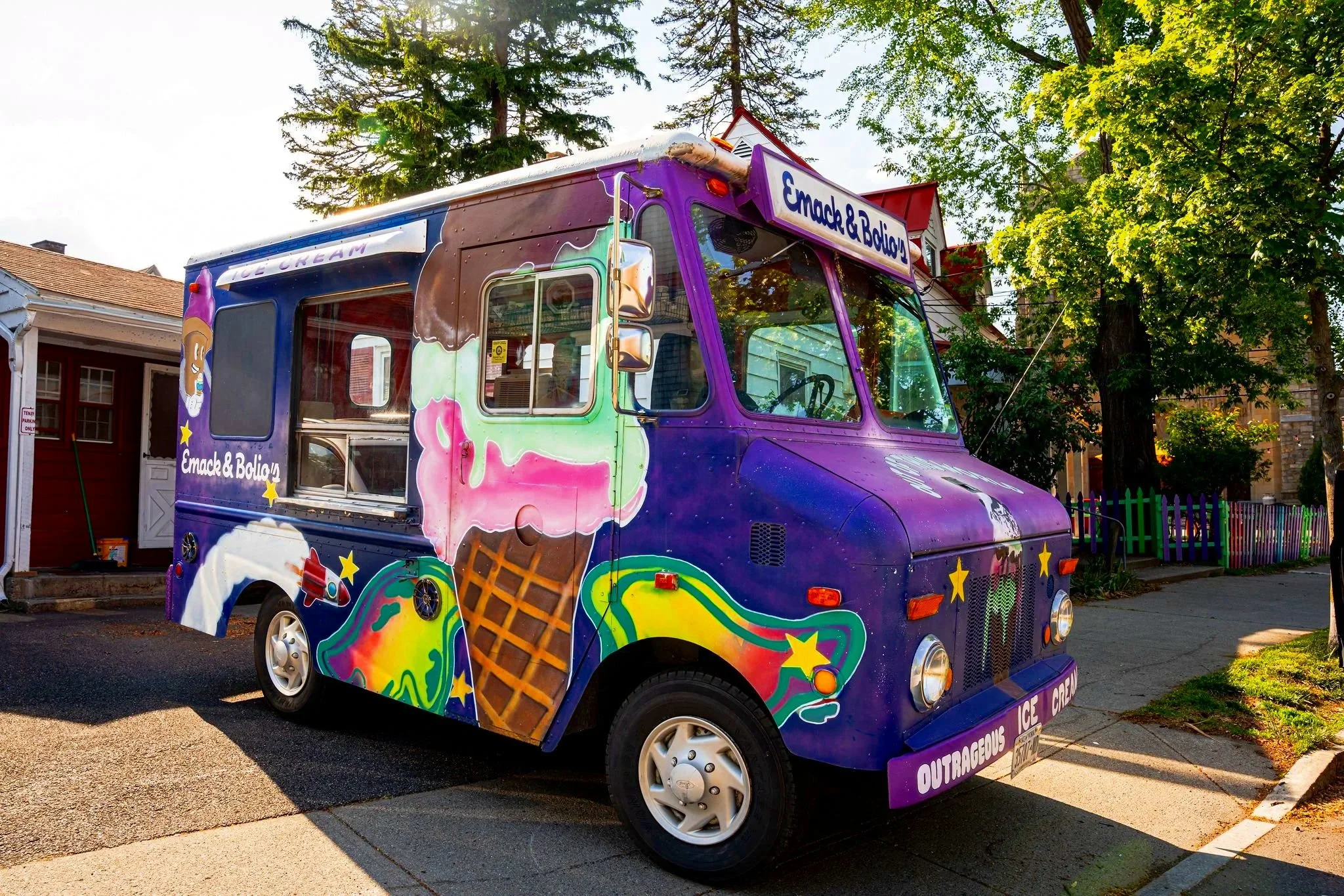 Colorful ice cream truck with images of ice cream cones and toppings parked on a street, trees and houses in the background.