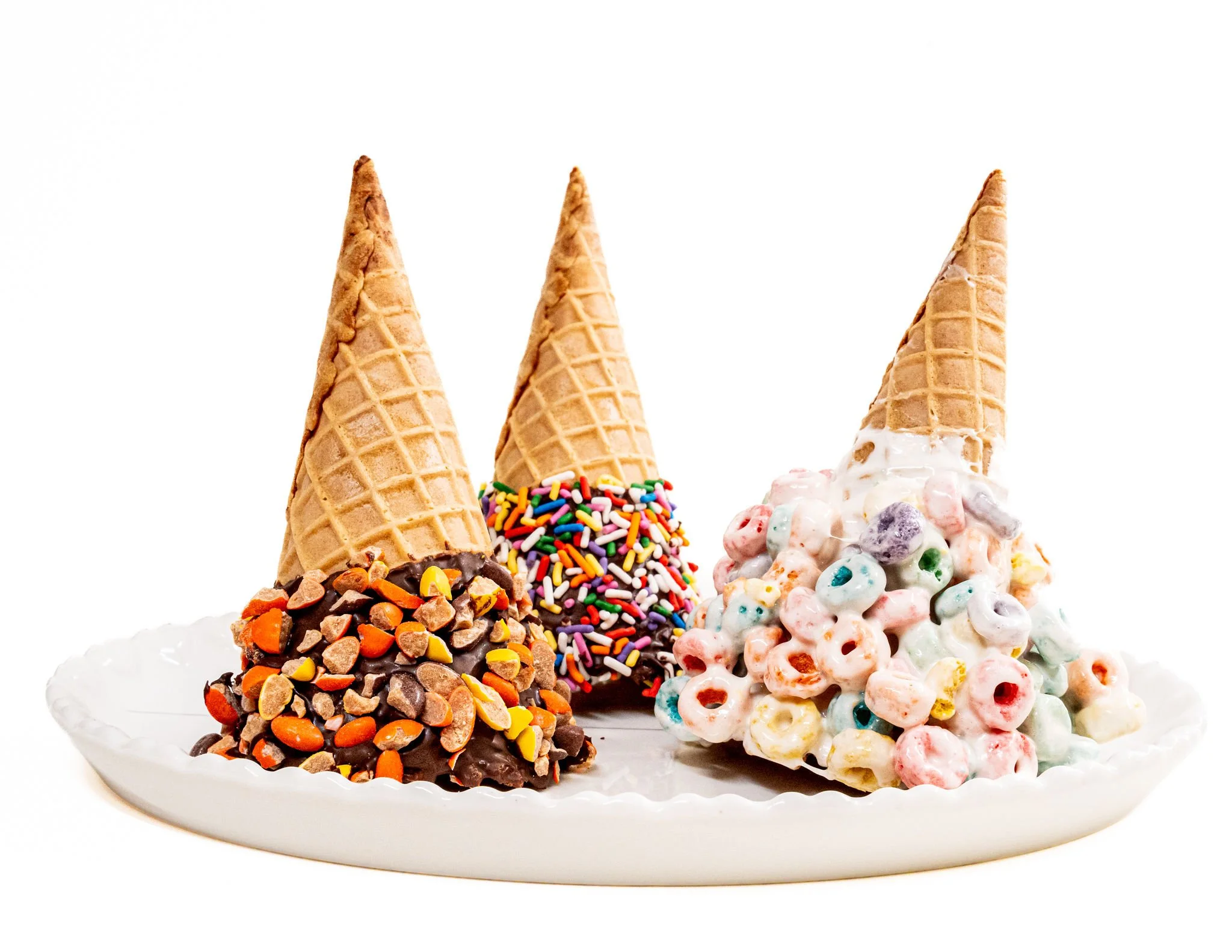 A white plate with three ice cream cones topped with colorful cereals and chocolate, served on a white surface.