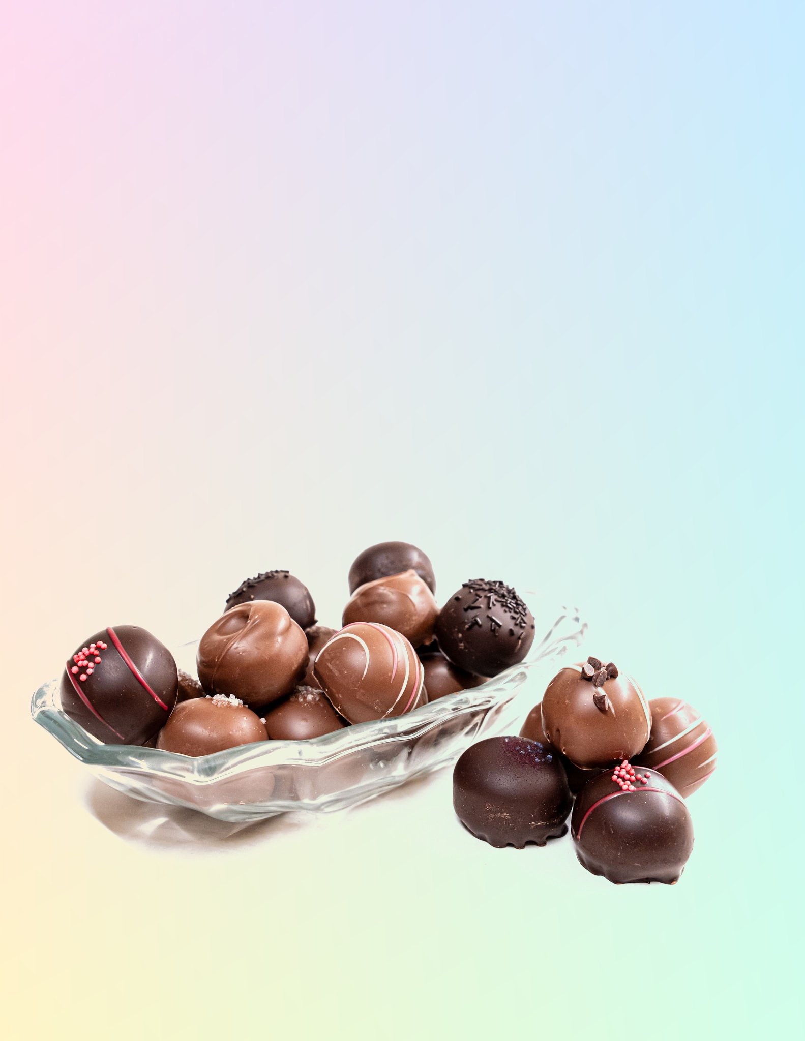 Assorted chocolate truffles in a glass bowl, decorated with sprinkles and drizzle, on a pastel gradient background.