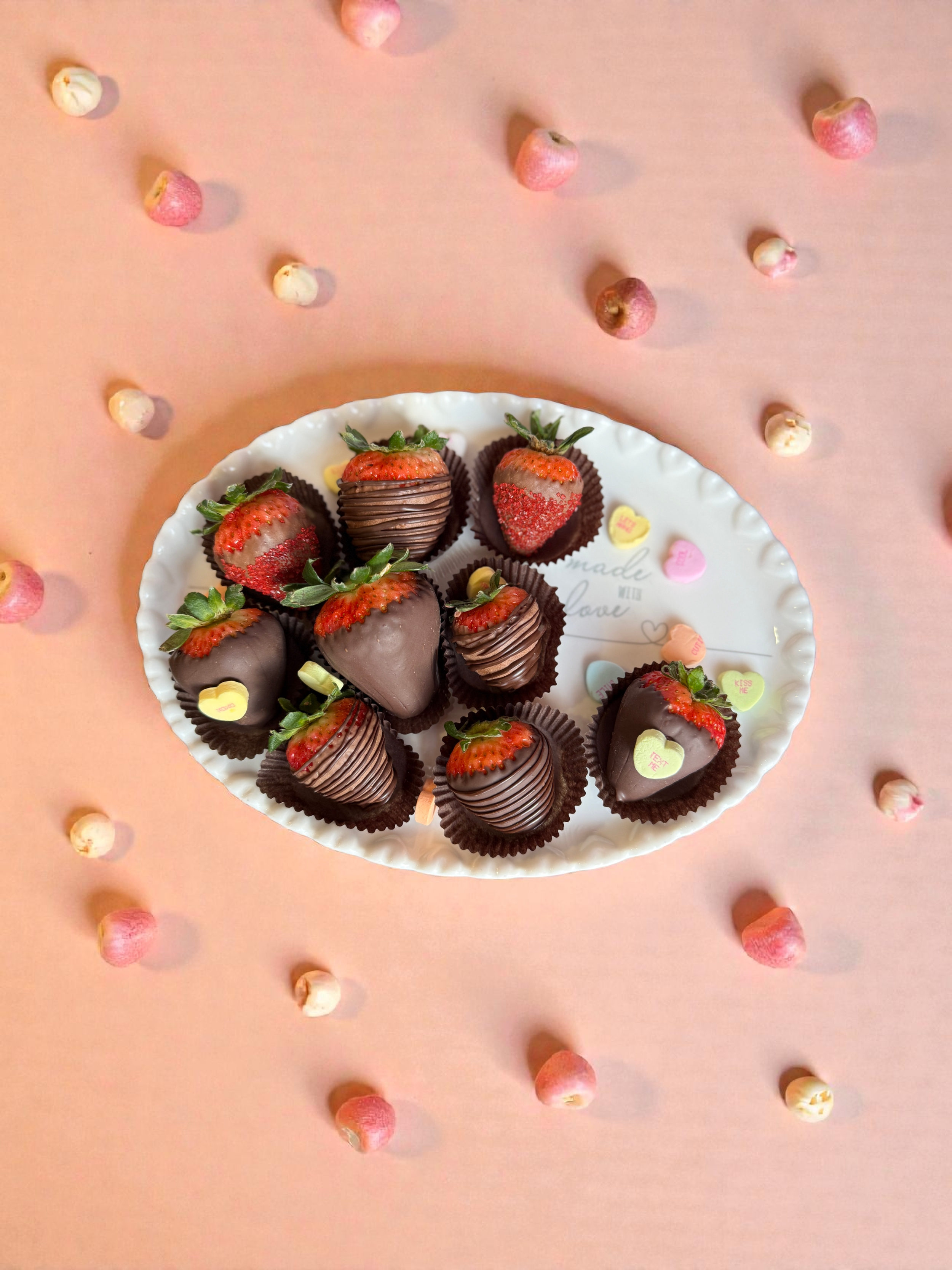 A white plate with chocolate-covered strawberries and heart-shaped candies on a pink background with scattered mini marshmallows.