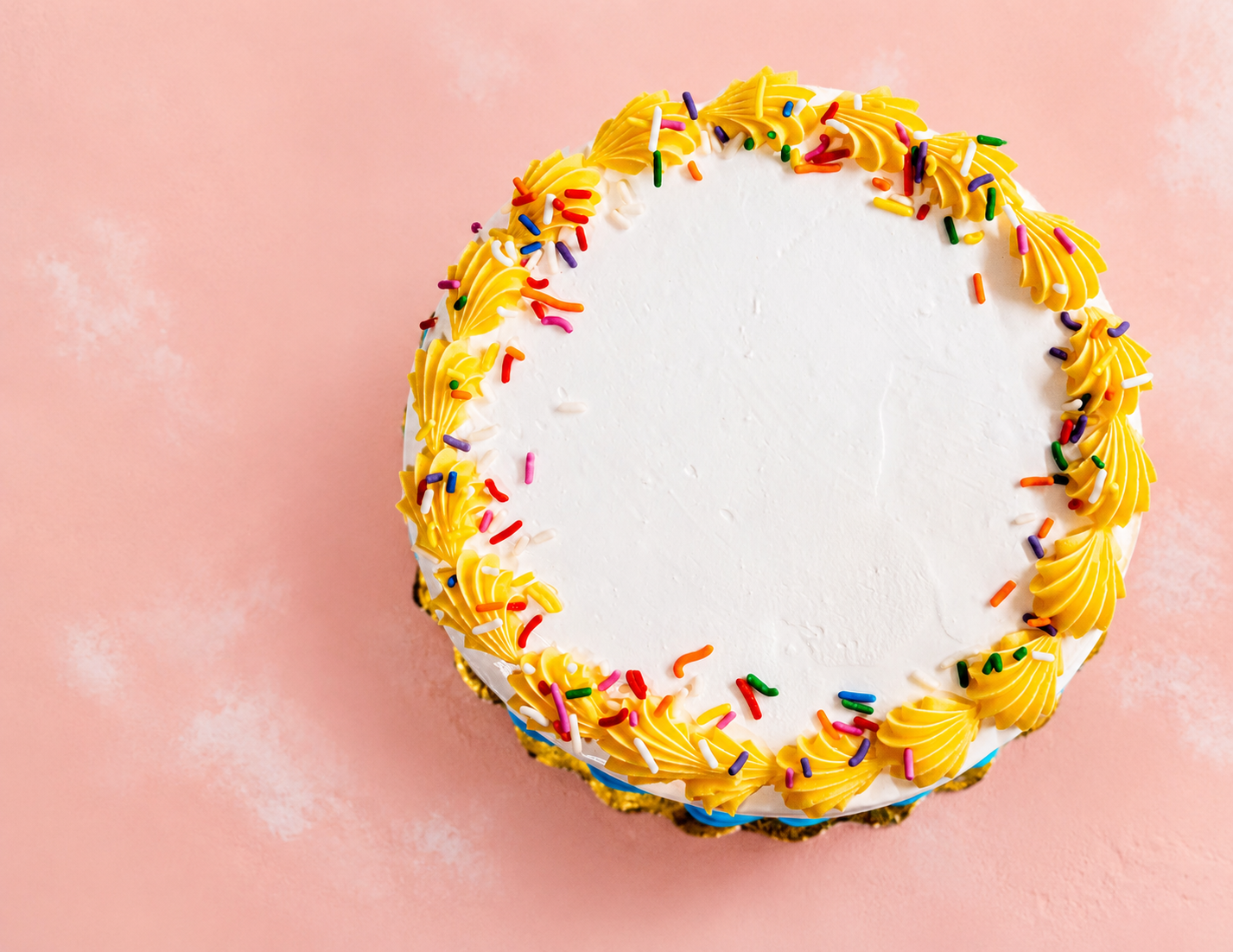 A round birthday cake with white frosting, decorated with yellow piped icing and colorful sprinkles on a pink background.