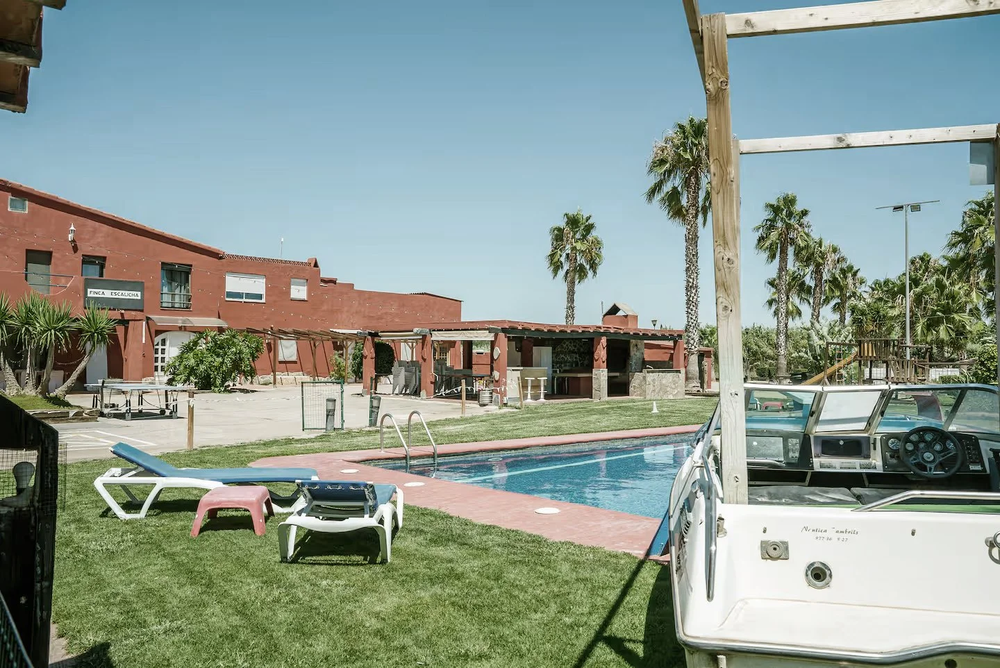 ride europe uk spain motocross holiday holiday's Resort pool area with lounge chairs, palm trees, and a boat docked at the side, under a clear blue sky.